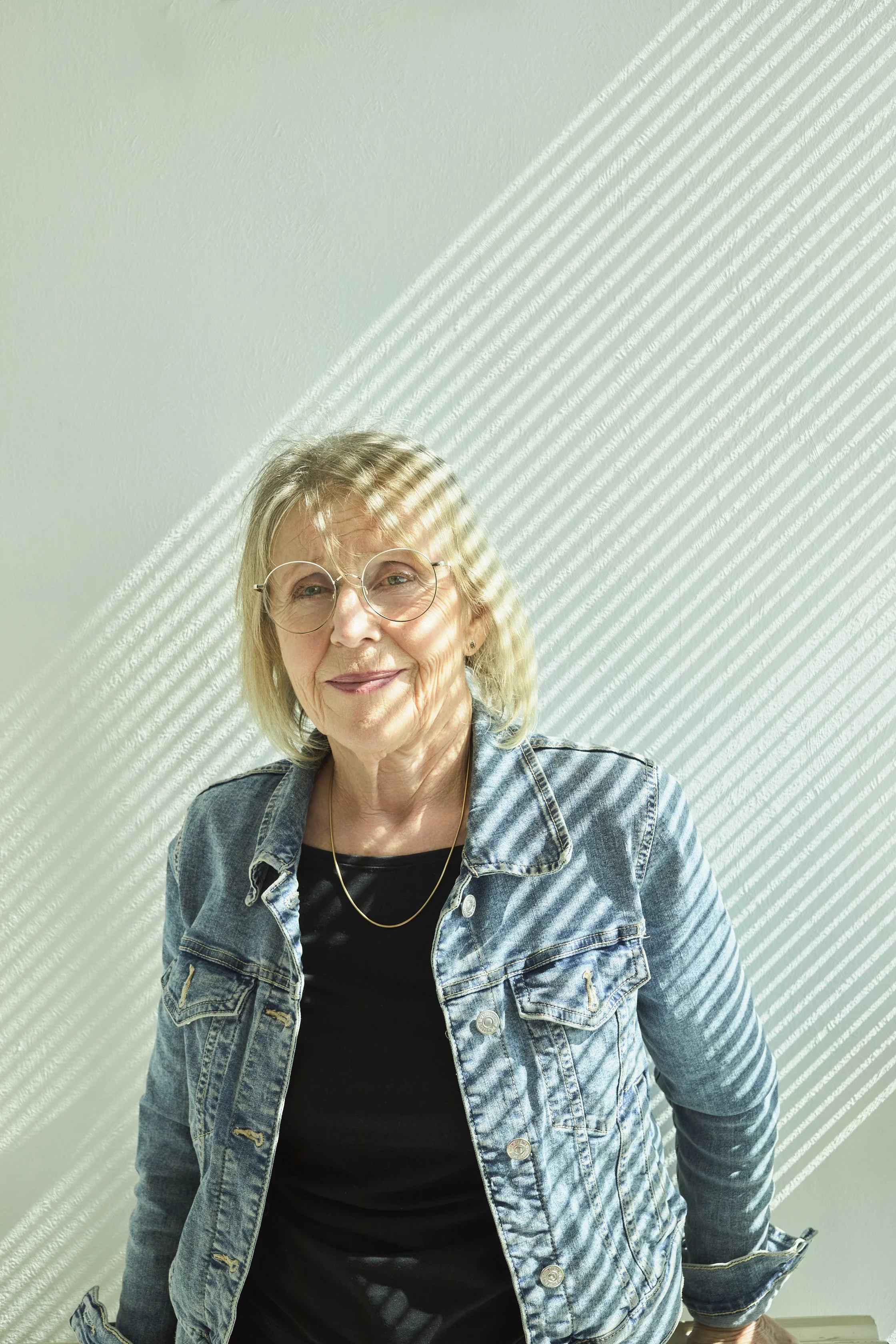 Une femme âgée portant des lunettes, une veste en jean, un t-shirt noir, et un collier, souriant, devant un mur blanc avec des rayures de lumière.