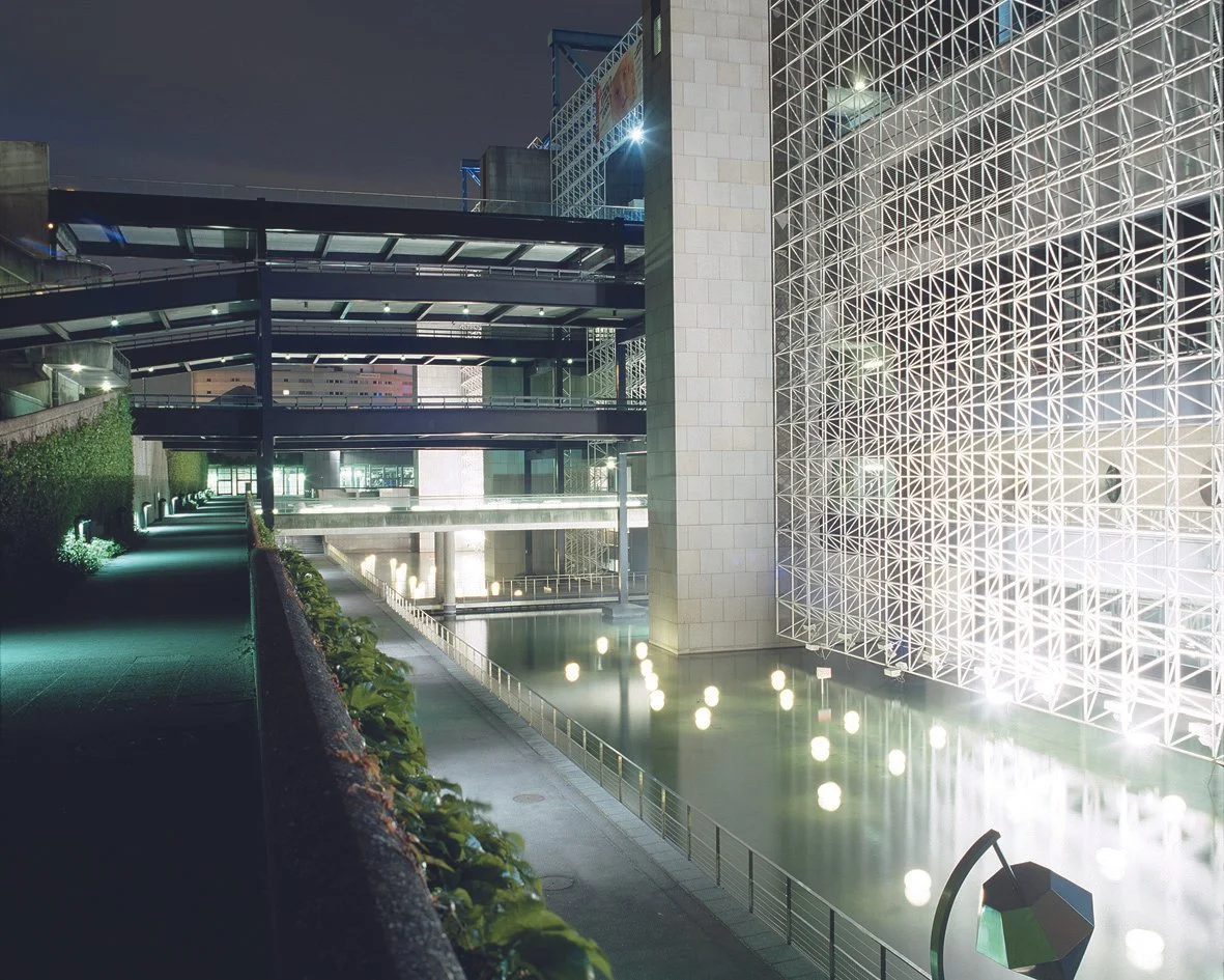 Vue nocturne d'un bâtiment moderne avec éclairage architectural, murs vitrés et une fontaine éclairée à l'eau calme, entourée de cheminements piétonniers.