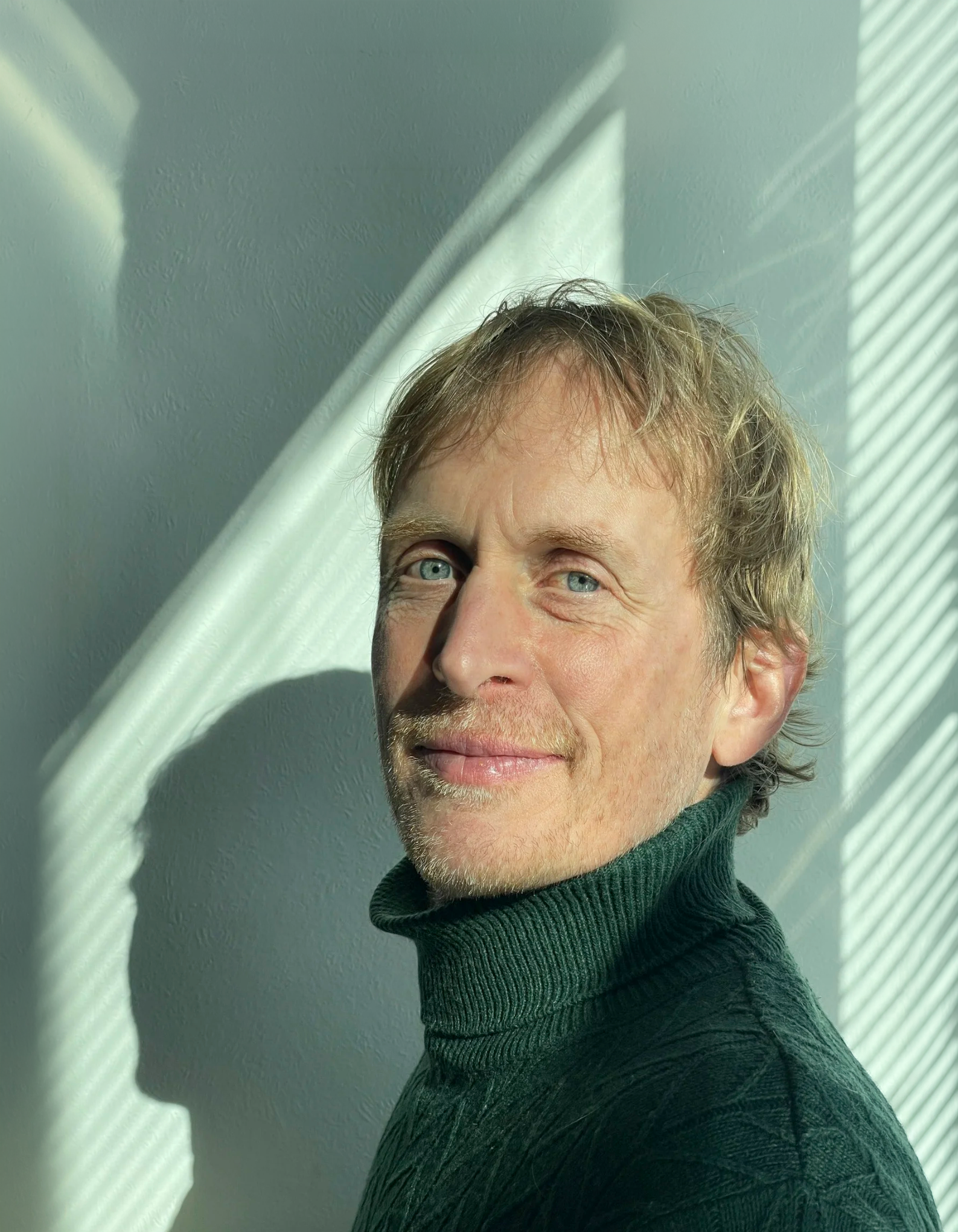 Un portrait d'un homme avec des cheveux bruns ondulés, des yeux bleus, portant un col roulé vert foncé, avec de la lumière naturelle créant des ombres et des motifs de rayures sur le mur derrière lui.