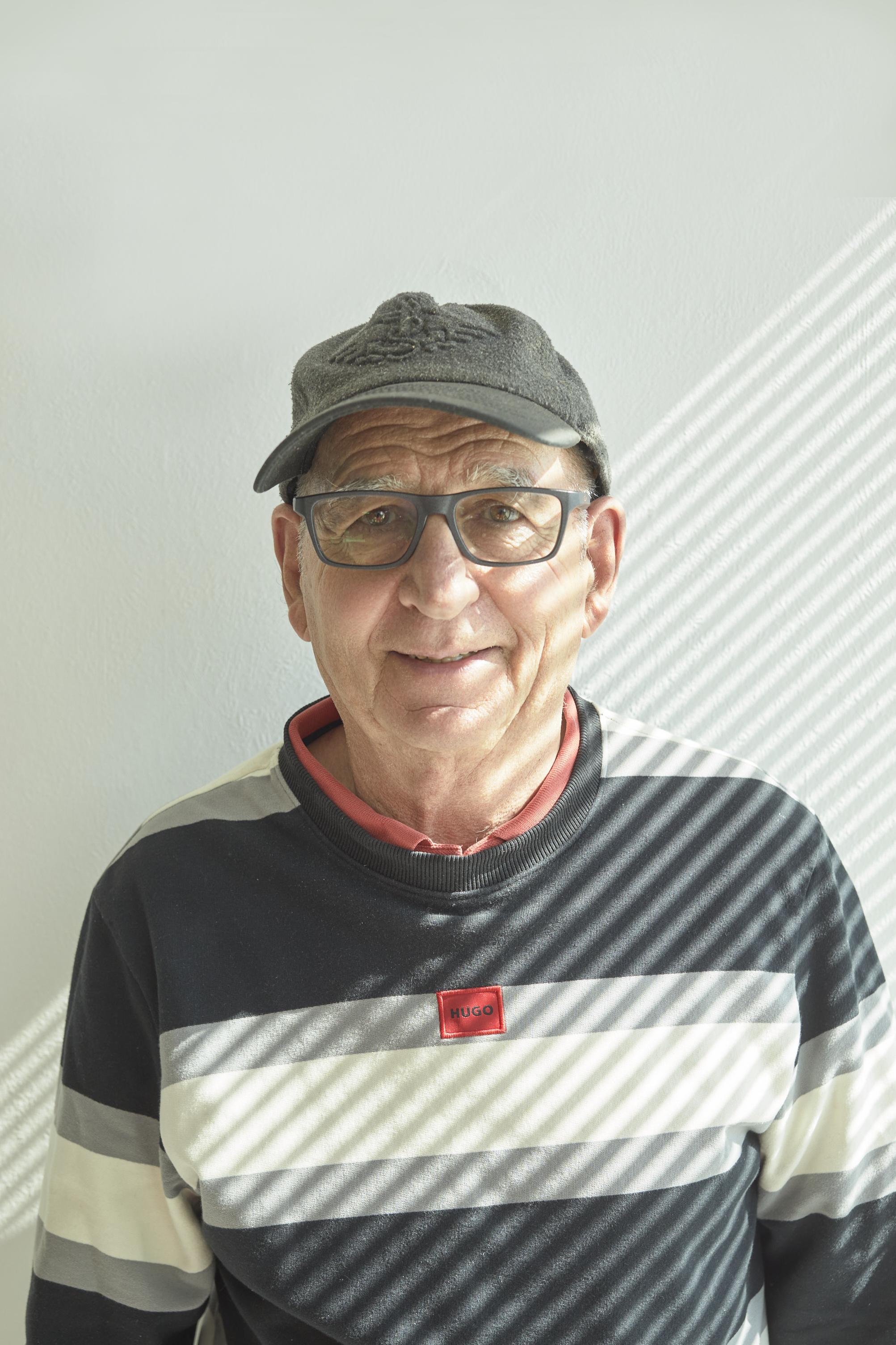 Un homme d'âge moyen porte une casquette grise, des lunettes et un sweat à rayures horizontales de couleurs noir, blanc et gris avec une étiquette rouge portant le nom 'HUGO'. Il sourit devant un mur blanc avec des rayons de lumière créant des lignes diagonales.