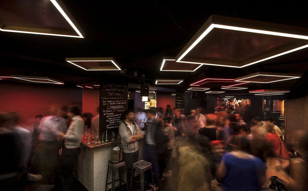 Un bar animé avec des gens qui socialisent, éclairage tamisé, panneaux noirs avec écriture blanche, plafond avec des lumières en forme de rectangles.