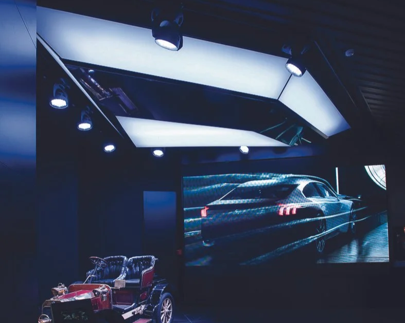 Une salle d'exposition avec une projection d'une voiture de luxe sur un grand écran, un petit véhicule électrique en face.