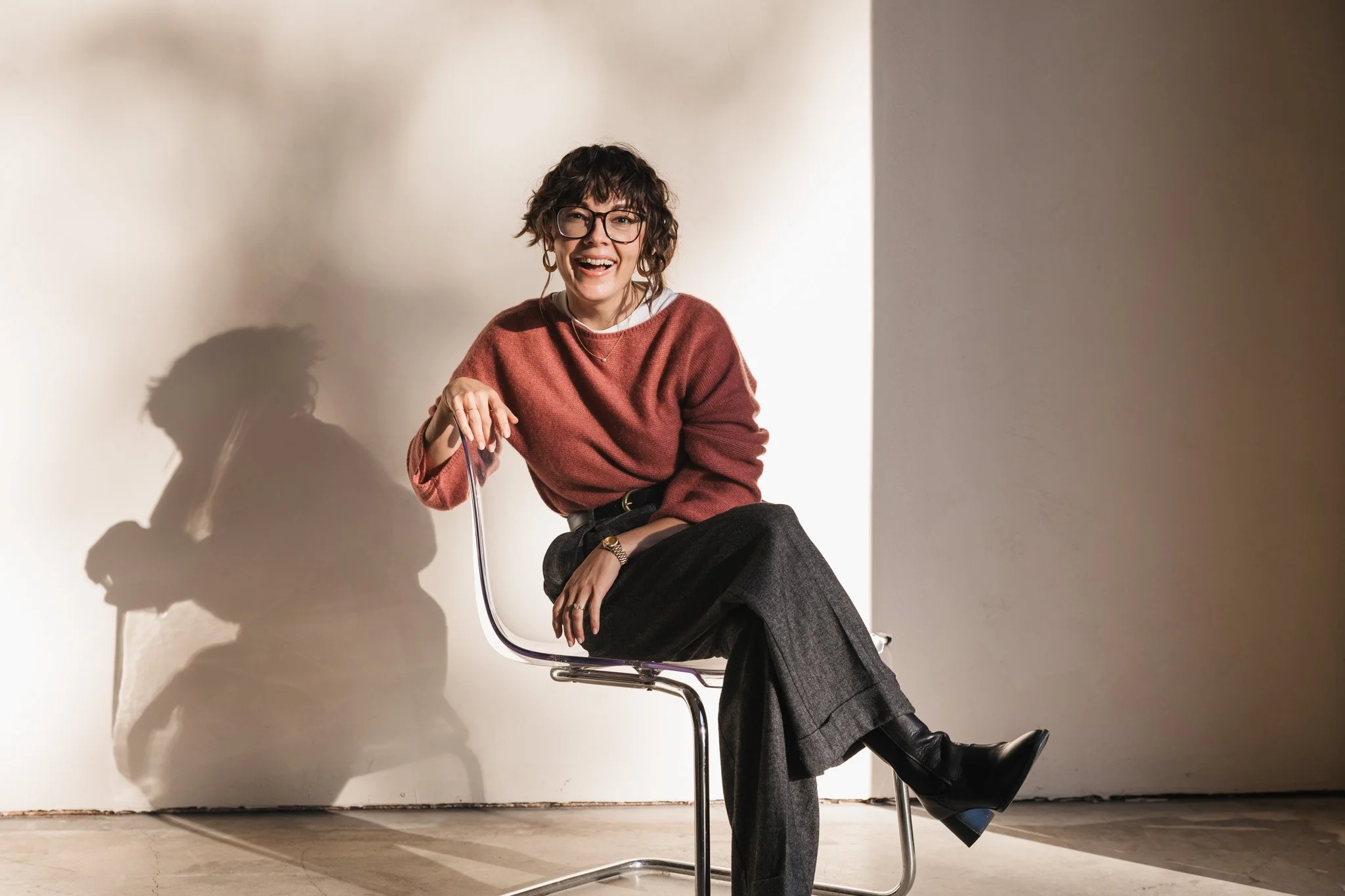 A woman with short curly hair and glasses, smiling while sitting on a modern chair, wearing a pink sweater and dark wide-leg trousers, against a plain wall and casting a shadow.