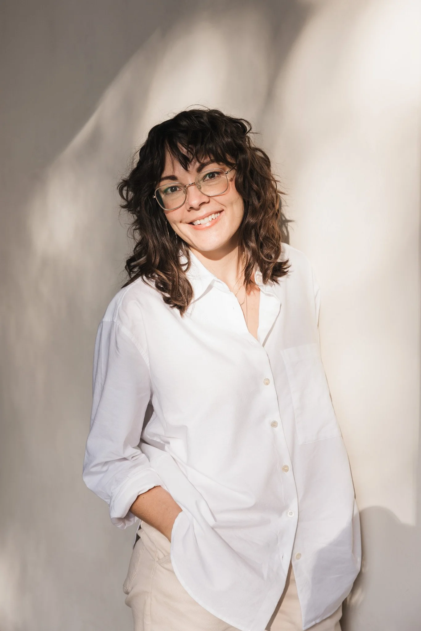Smiling woman with glasses wearing a white shirt and beige pants, standing with hands in pockets against a blurred background.