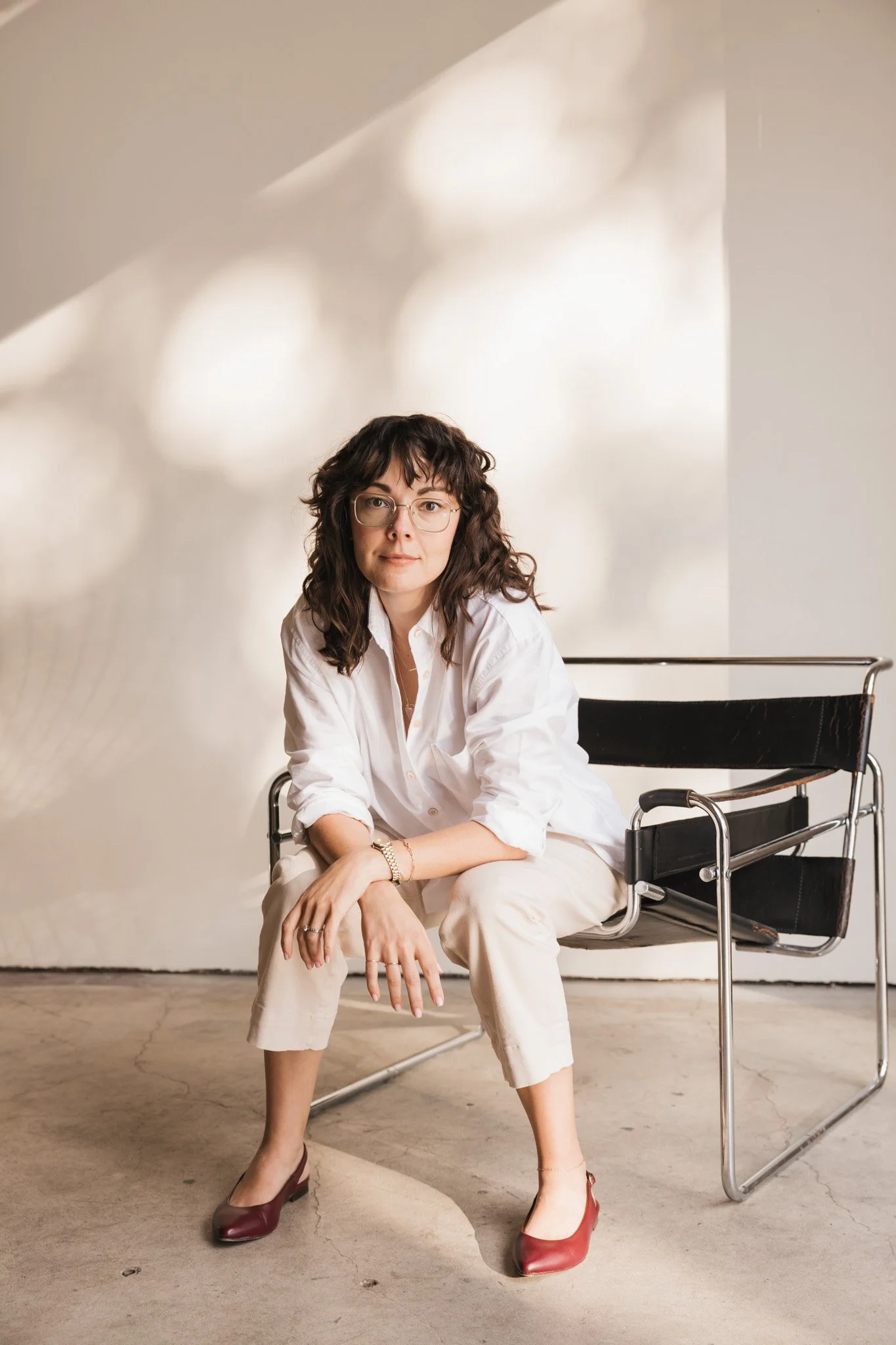 A woman with dark, curly hair and glasses sitting on a modern black and chrome chair in a minimalist room with a concrete floor and soft natural light. She wears a white shirt, beige pants, and red shoes.