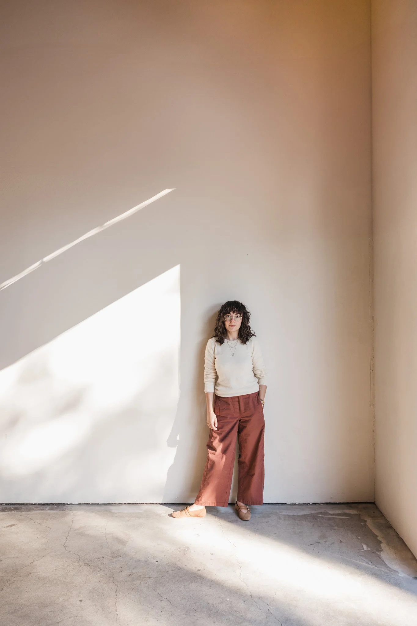 Woman with dark curly hair and glasses standing against a beige wall with natural sunlight casting shadows on the wall and floor.