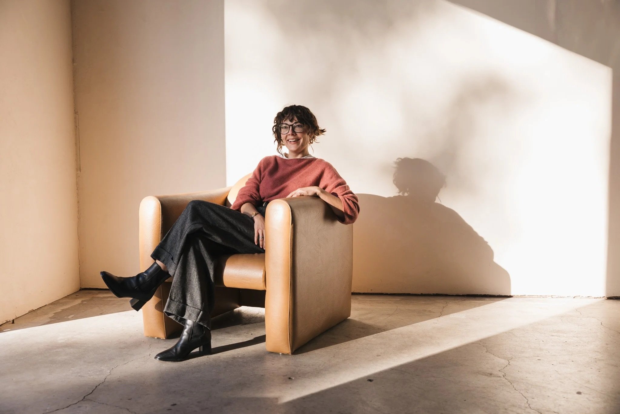 A woman with glasses and curly hair sitting on a tan armchair, smiling. She is wearing a pink sweater, black pants, and black boots. The setting is minimalist with natural light casting a shadow on the wall behind her.