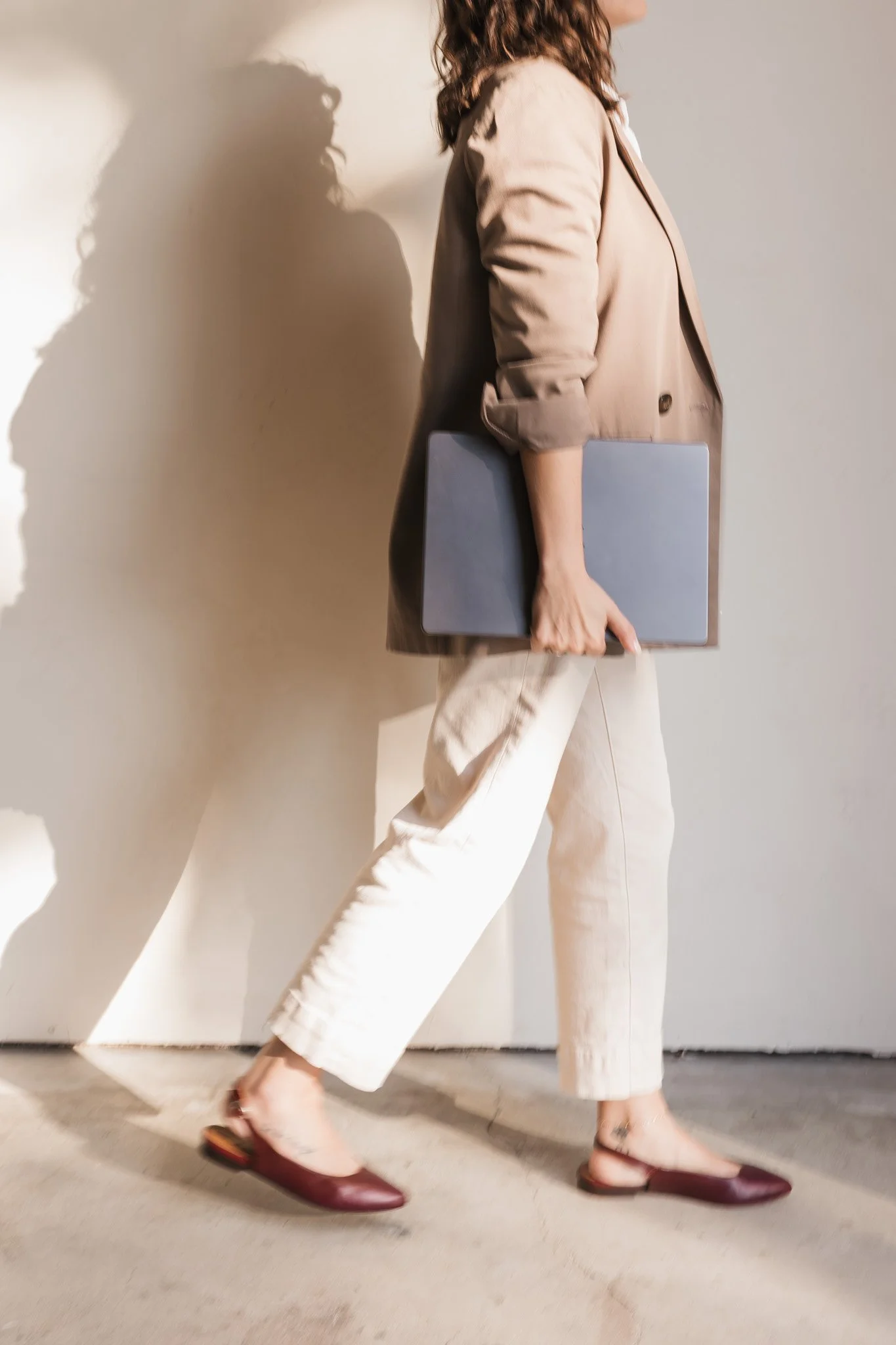 A woman walking with a laptop in her hand, wearing beige pants, a beige blazer, and burgundy flats, casting a shadow on the wall.