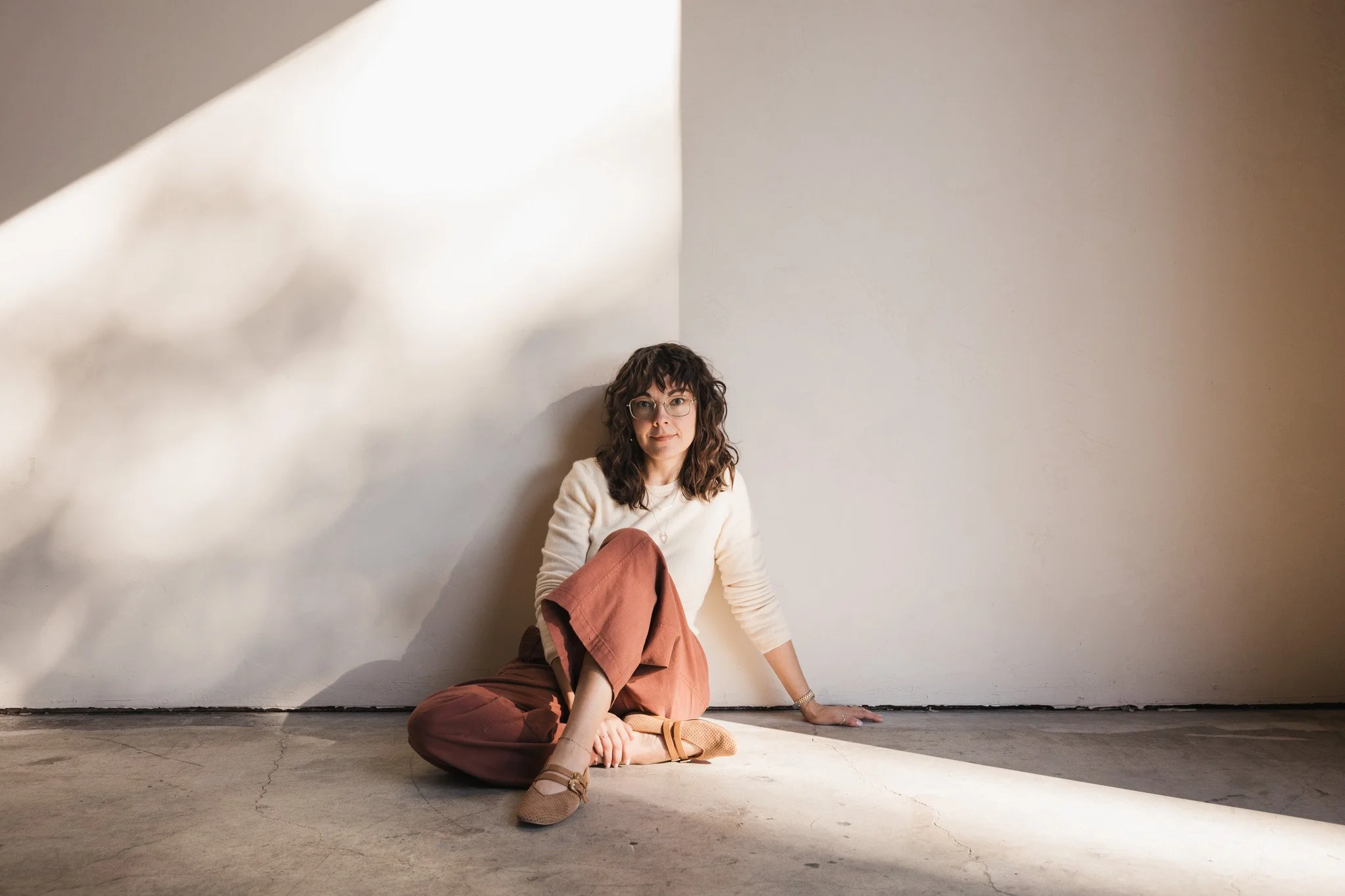 A woman with curly hair and glasses sitting on a concrete floor, leaning against a white wall with sunlight creating a shadow on the wall.