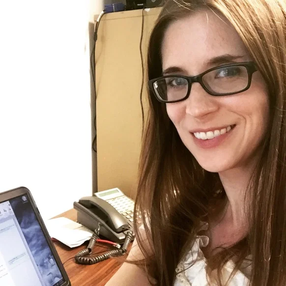 Emily is wearing glasses and sitting at a desk in front of a laptop.