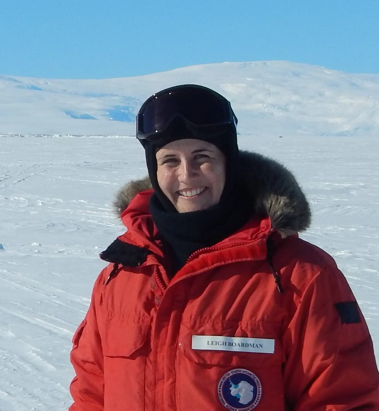 Leigh is wearing heavy winder gear such as a red jacket, black hat and snow goggles, while standing in front of a snowy backdrop with blue skies.