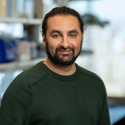 Mihir wearing a green sweater with a blurred background of a lab