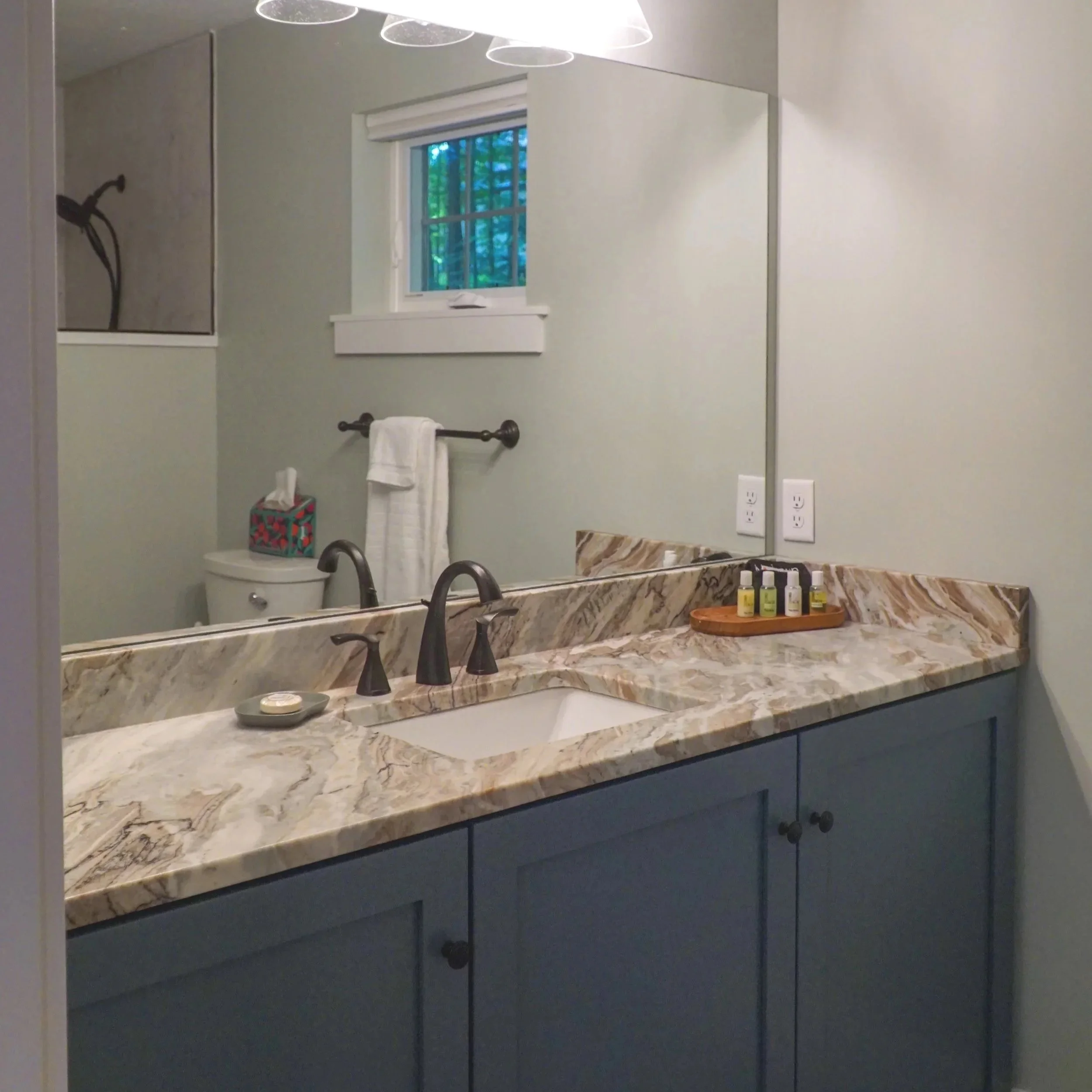 A bathroom vanity with a marble countertop, blue cabinet doors, a large mirror, and a set of small toiletries on a wooden tray, with a window and towel rack reflected in the mirror.