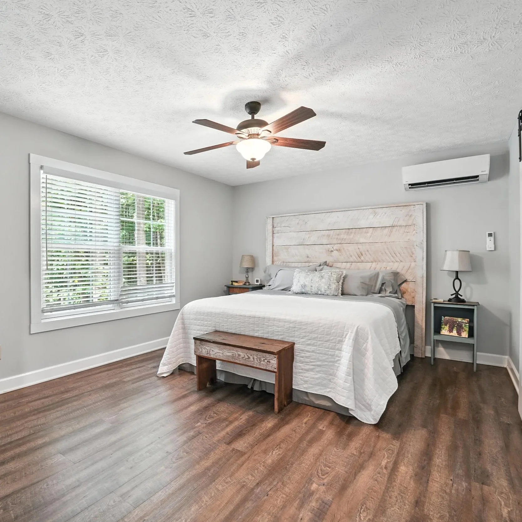 A bedroom with a large bed, white bedding, wooden headboard, and two nightstands with lamps. There is a window with blinds, a ceiling fan, a wall-mounted TV, and an air conditioner.
