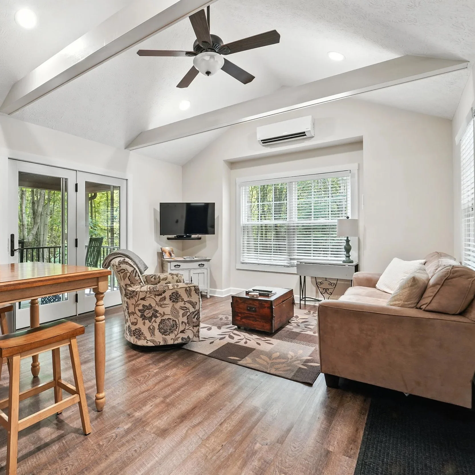 Living room with a beige couch, an armchair, a TV mounted on the wall, a window with blinds, and a glass sliding door leading to a balcony surrounded by trees.