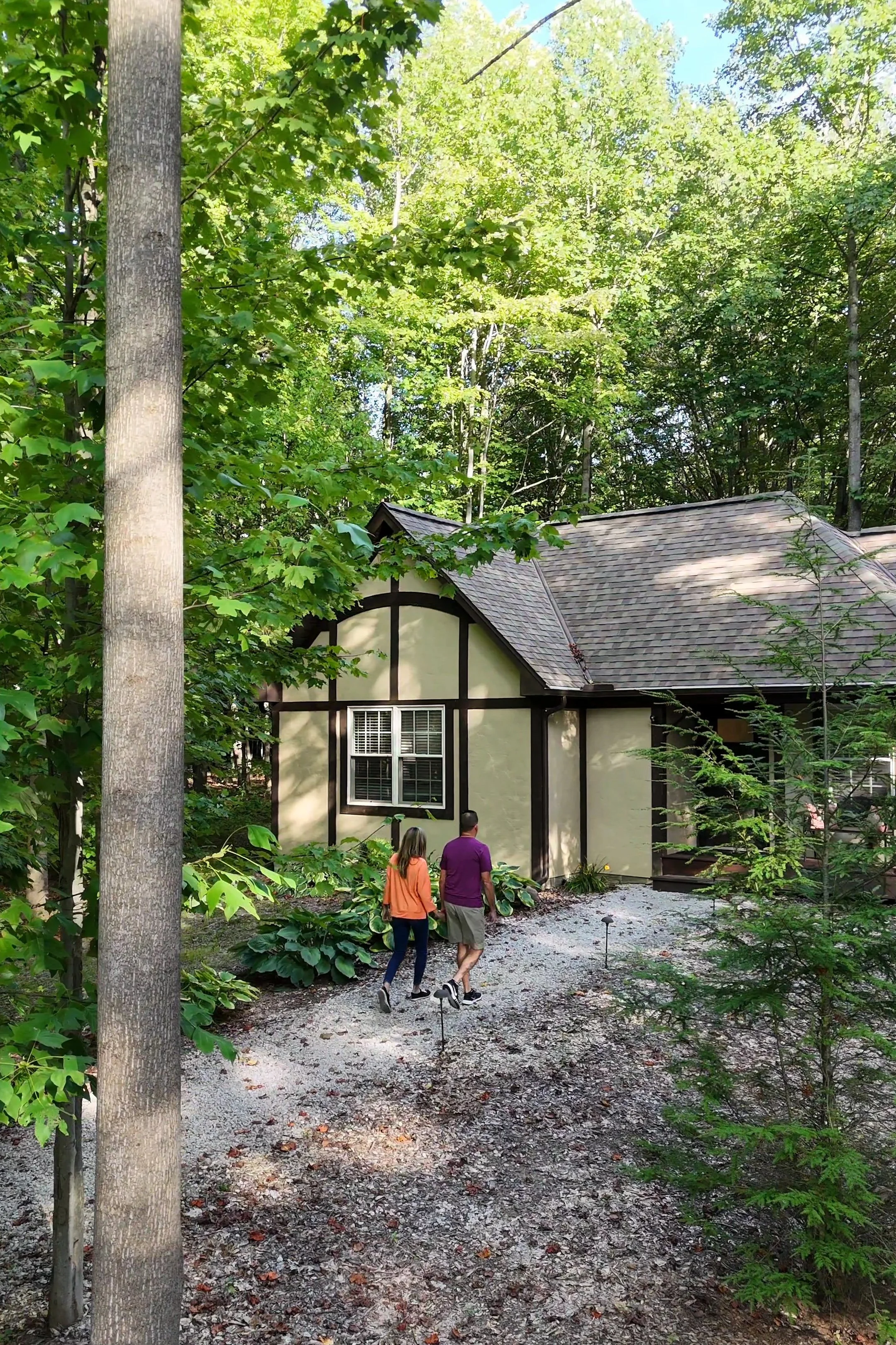 A couple of people walking on a gravel path in a wooded area near a house with a gabled roof and windows, surrounded by green trees and plants.
