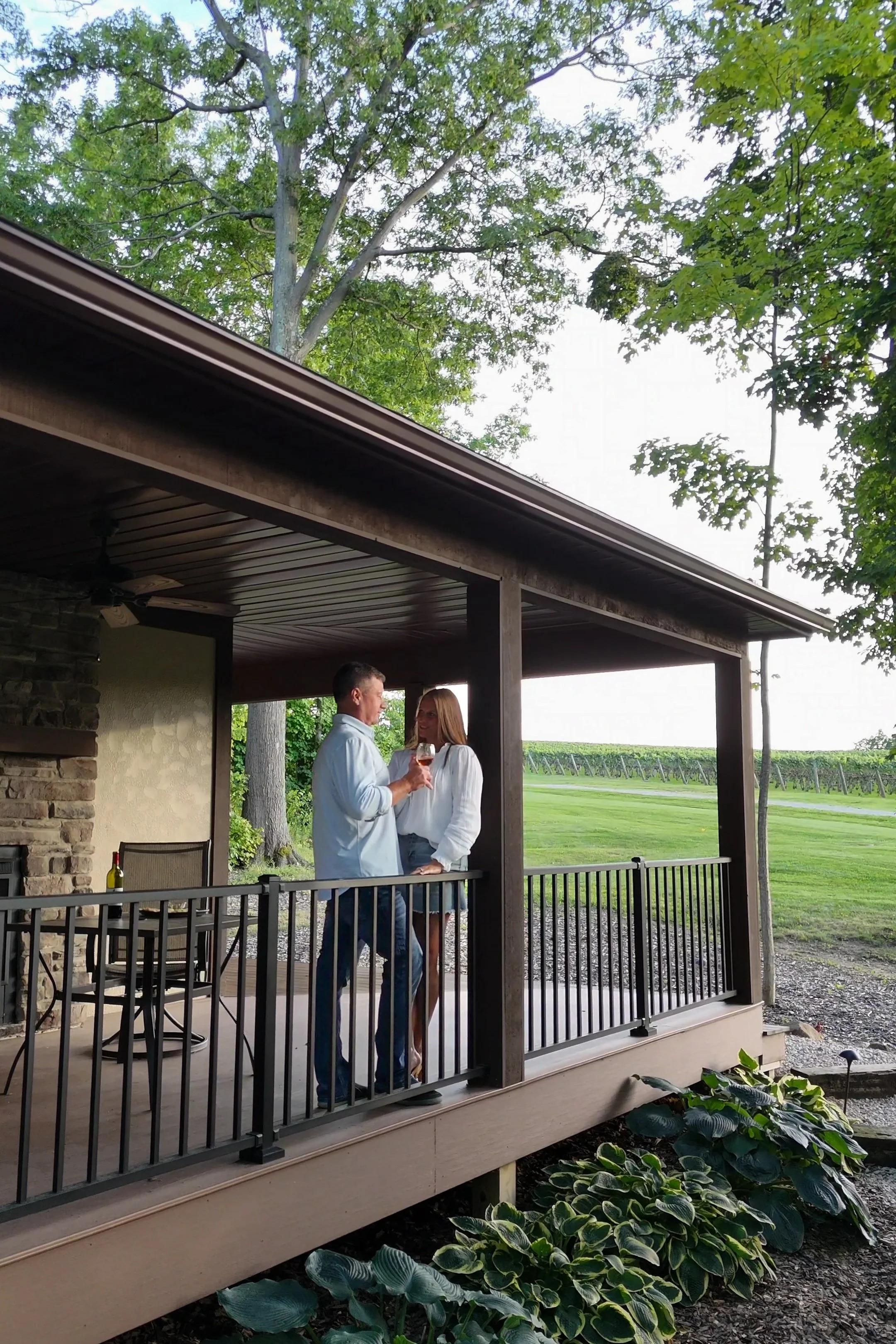 A man and a woman are standing on a porch under a roof, holding a glass of wine and having a conversation. They are outdoors in a rural setting with green grass, trees, and a vineyard in the background.