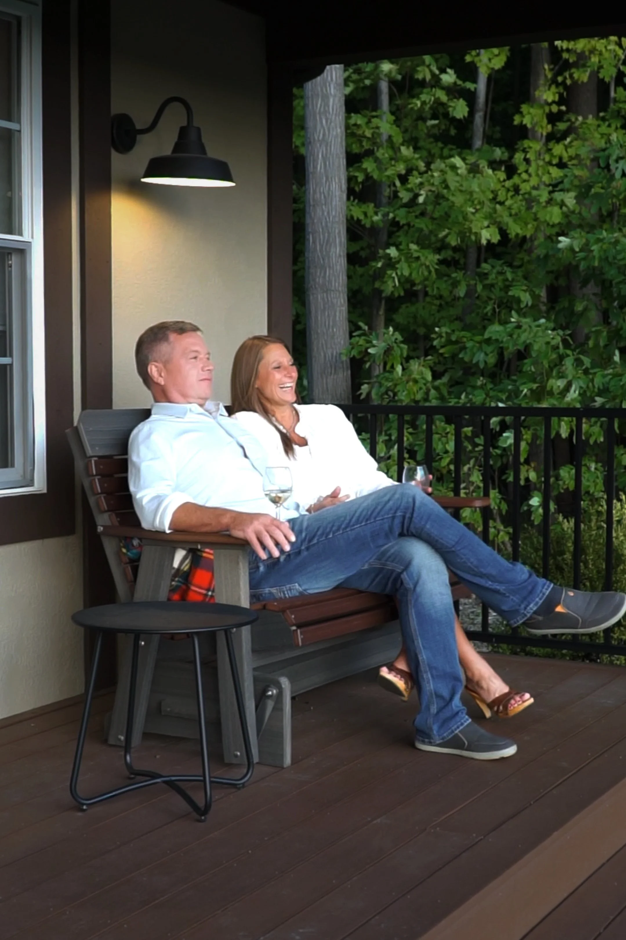 A couple is sitting on a porch bench, enjoying drinks, with a background of green trees and an outdoor wall with a black wall light.  Chardonnay Cottage in Ohio Wine Country.