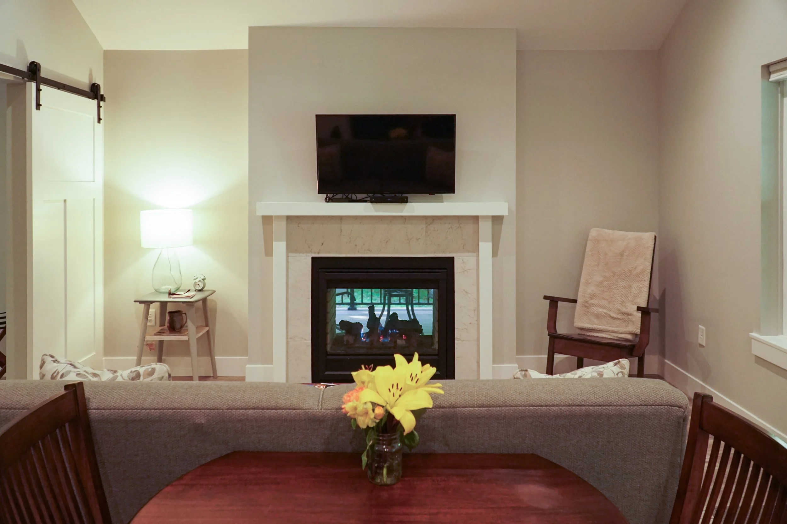 Living room with a fireplace, a mounted TV, a table lamp on a side table, a chair with a blanket, and a dining table with a vase of yellow flowers.