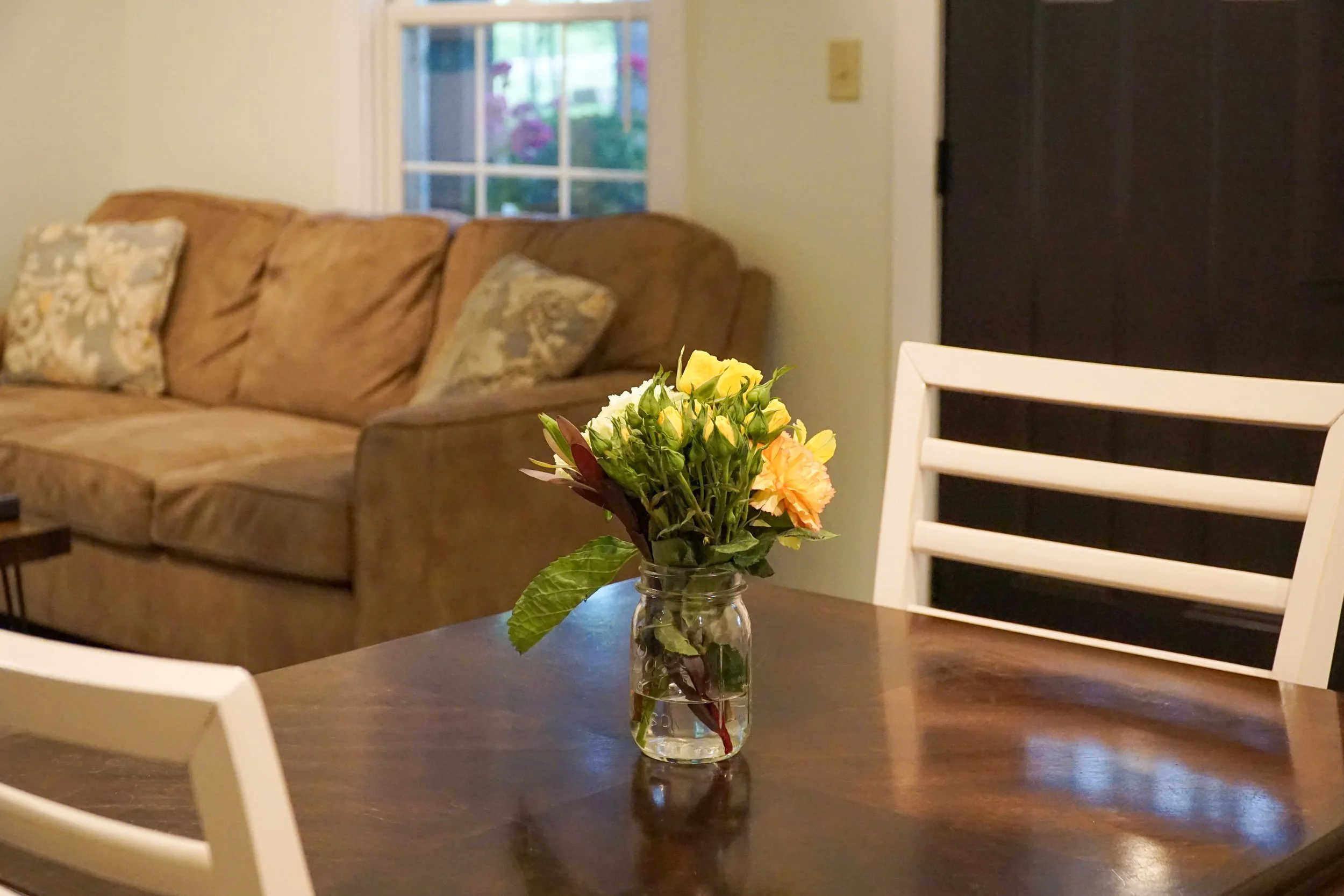 A bouquet of yellow, white, and peach flowers in a small glass jar on a dark wooden dining table.