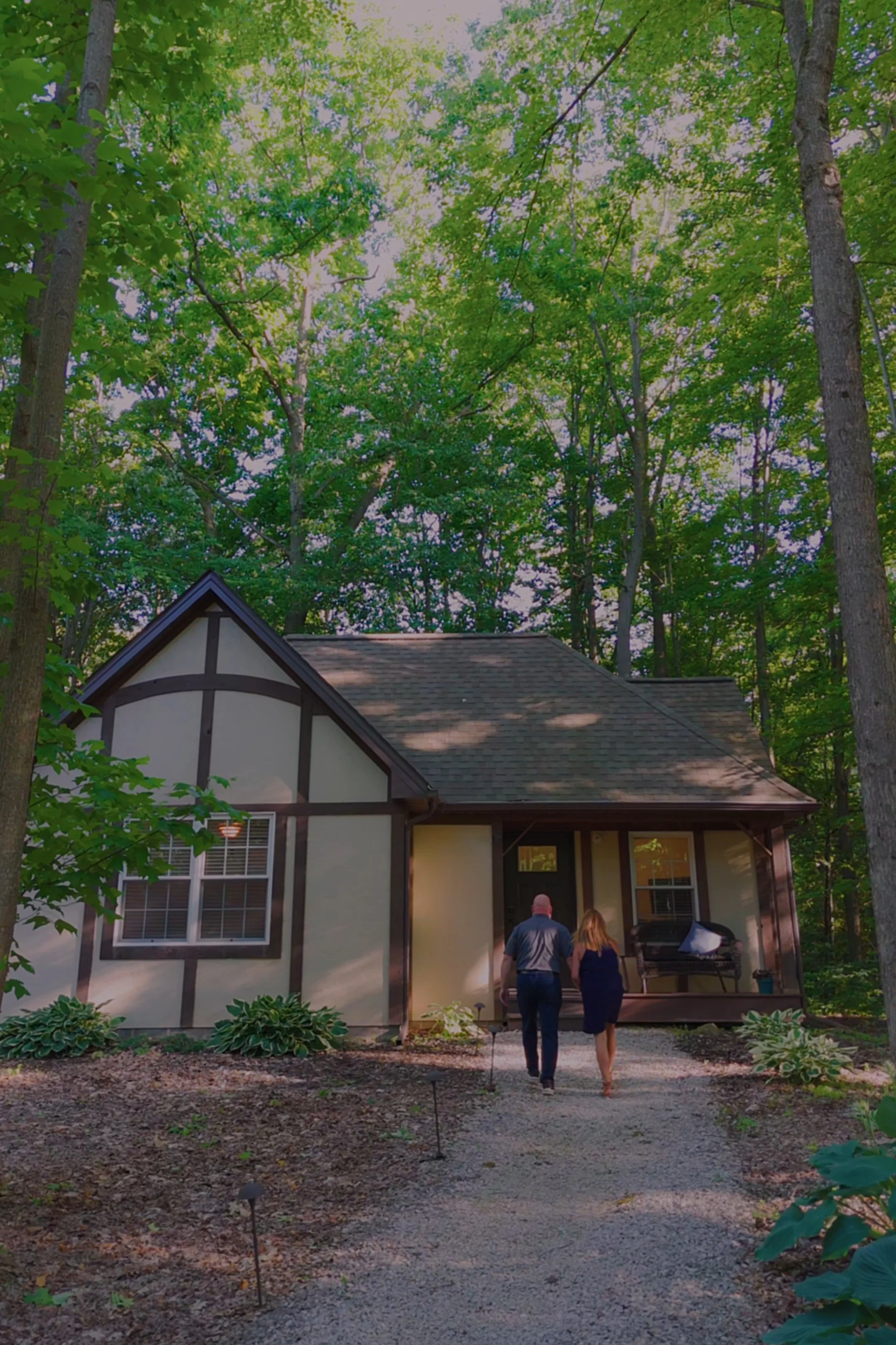 A man and woman walking towards the back door of a house in a wooded backyard at dusk.