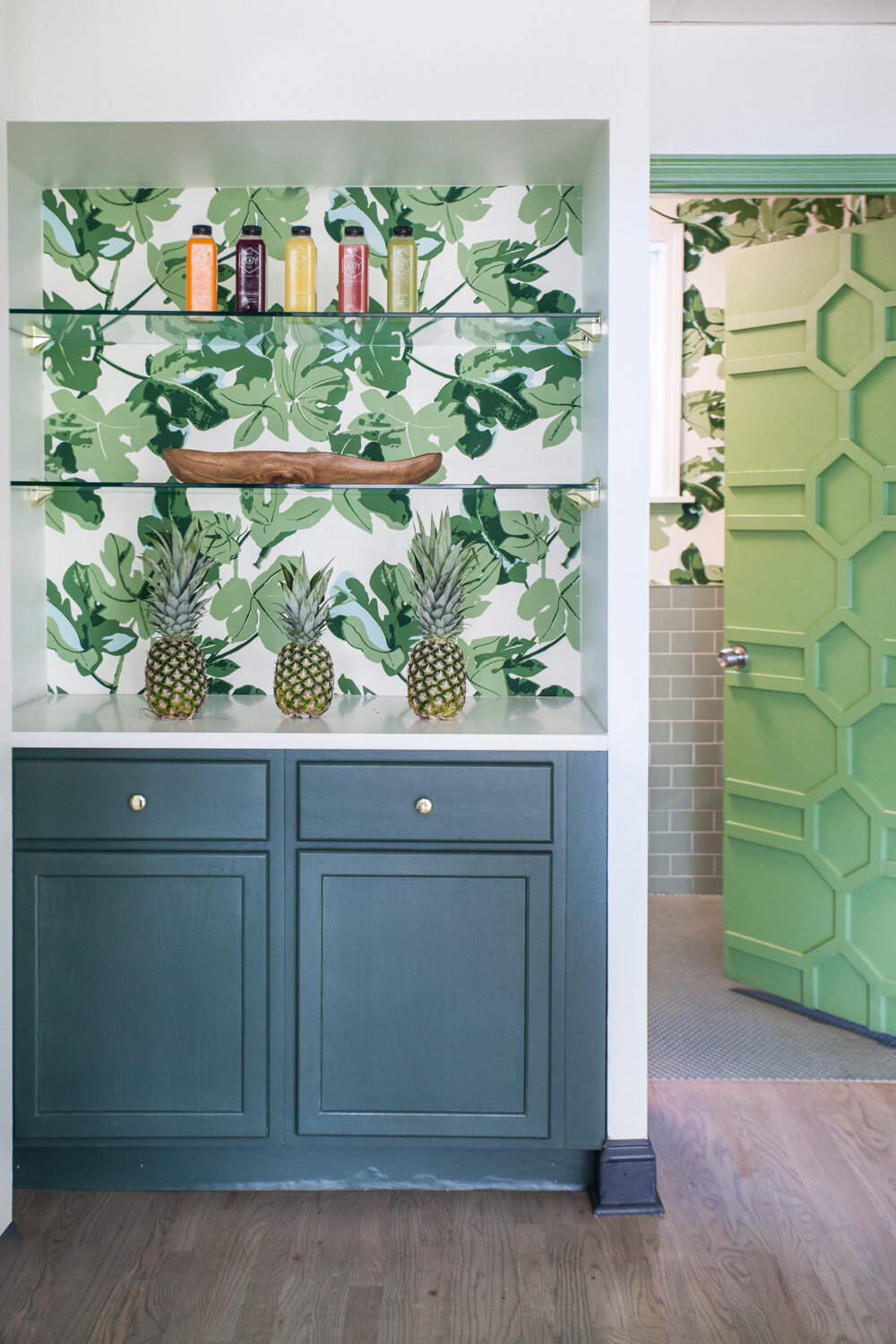 A welcoming Peter Dunham Fig Leaf Wallpaper, with glass shelving above a semi-custom cabinet painted in Benjamin Moore Dakota Shadow (448). Custom millwork door leads to a restroom.