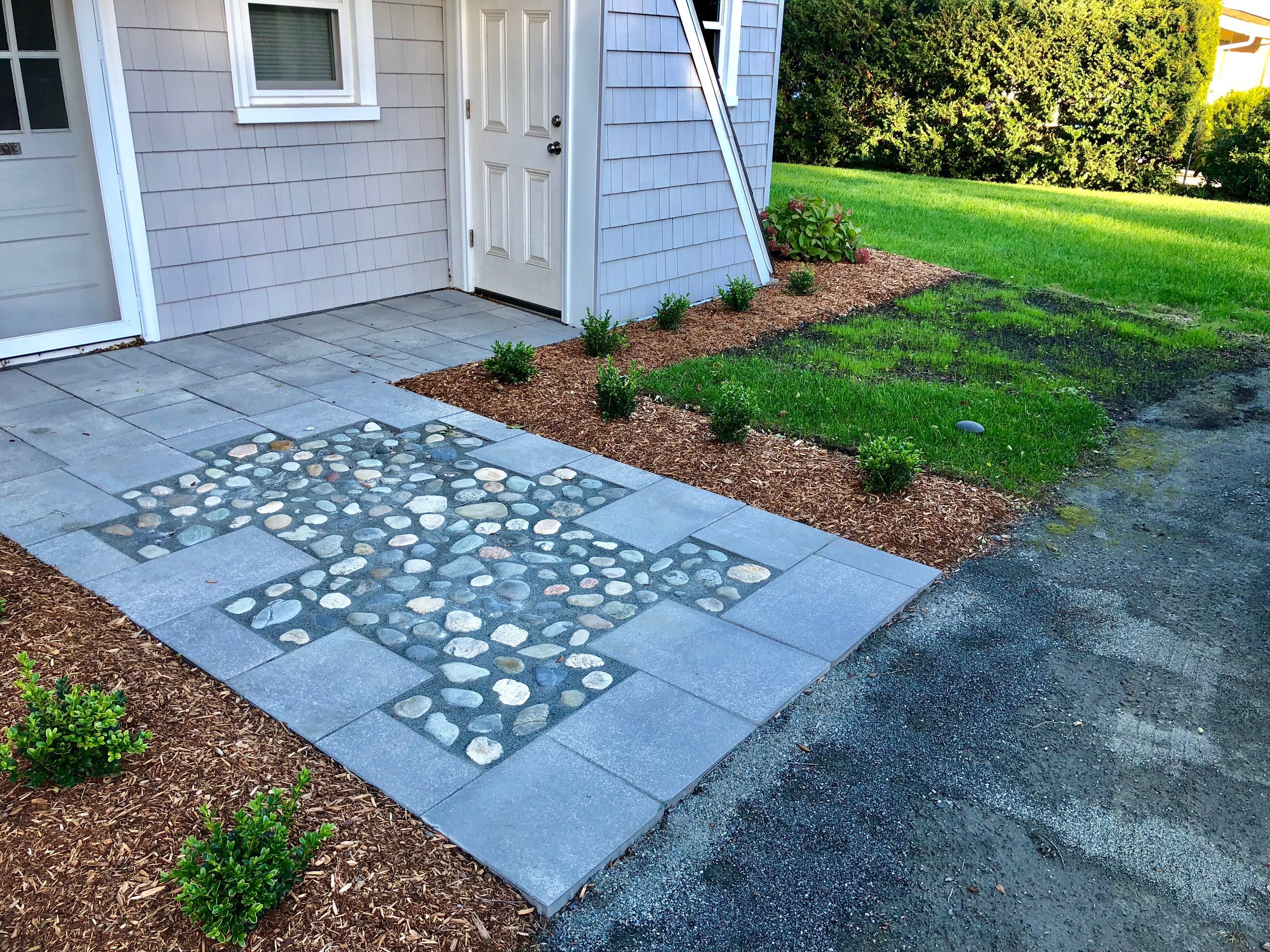 The new front hardscaping features a paver walkway framing an inset area composed of local beach stones. New plantings include boxwoods which will form a low hedge to frame the new path.