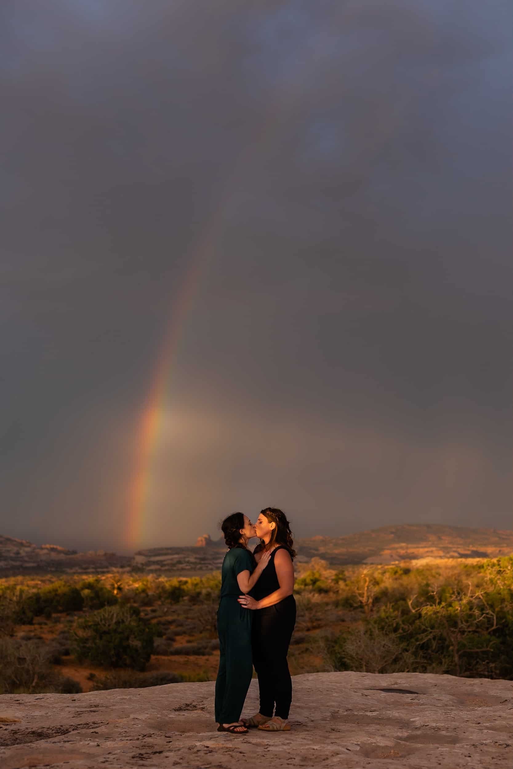 Lesbian couple kiss in front of a rainbow during their Moab engagement photoshoot
