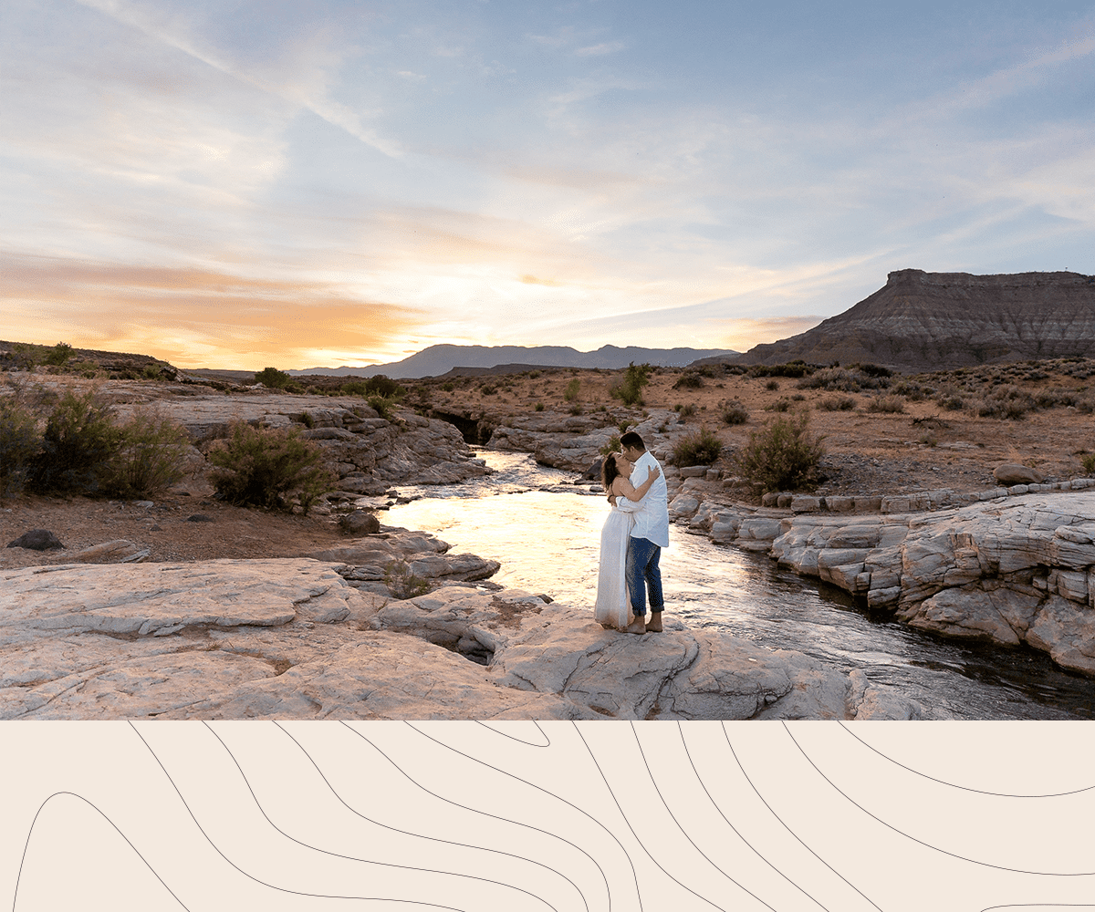 Couple embrace for a while landscape photo of them in front of a river and a sunset in Southern Utah Adventure Session Photographer