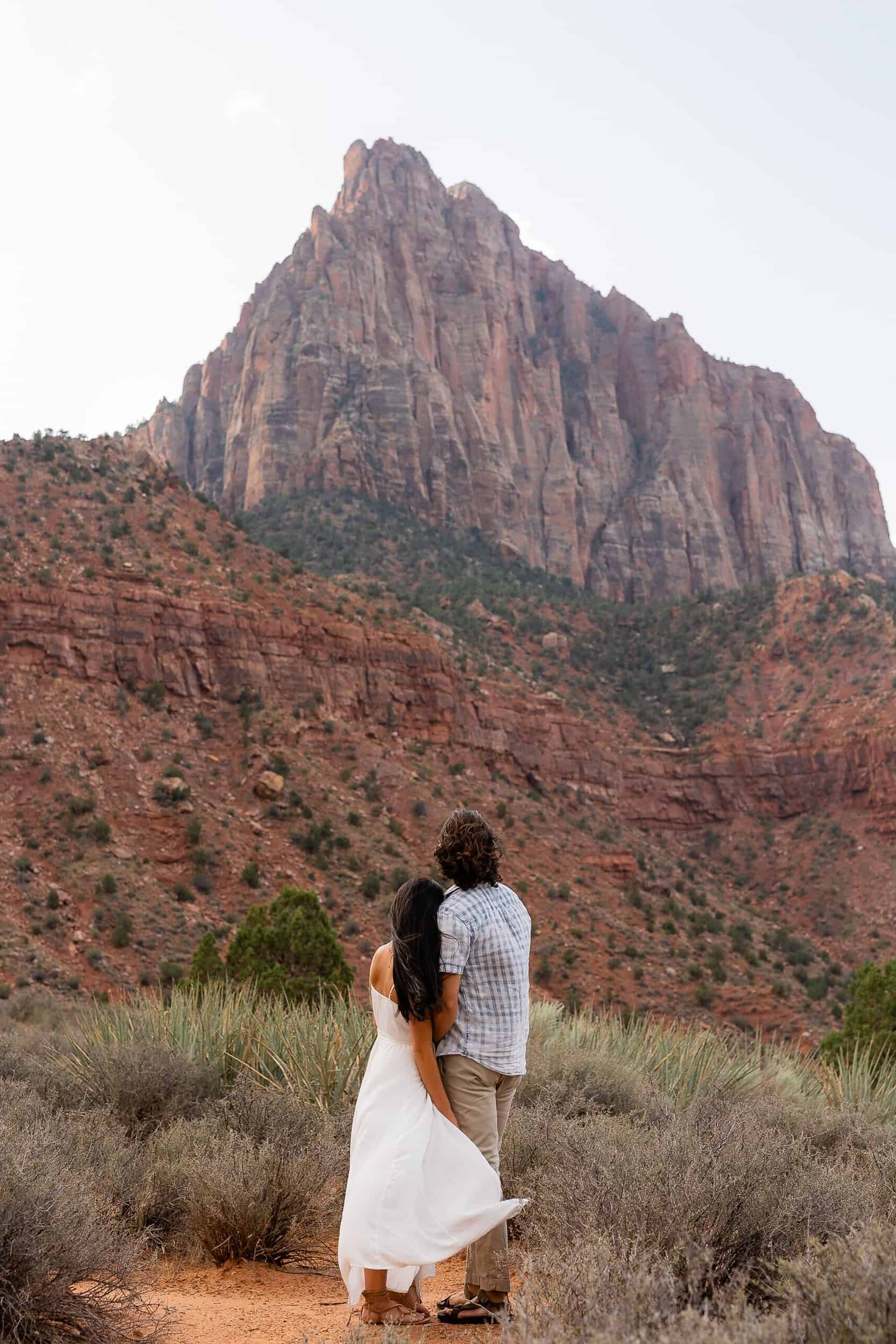 Couple hold each other facing a mountain in the distance during their adventure engagement photoshoot in Zion National Park, UT