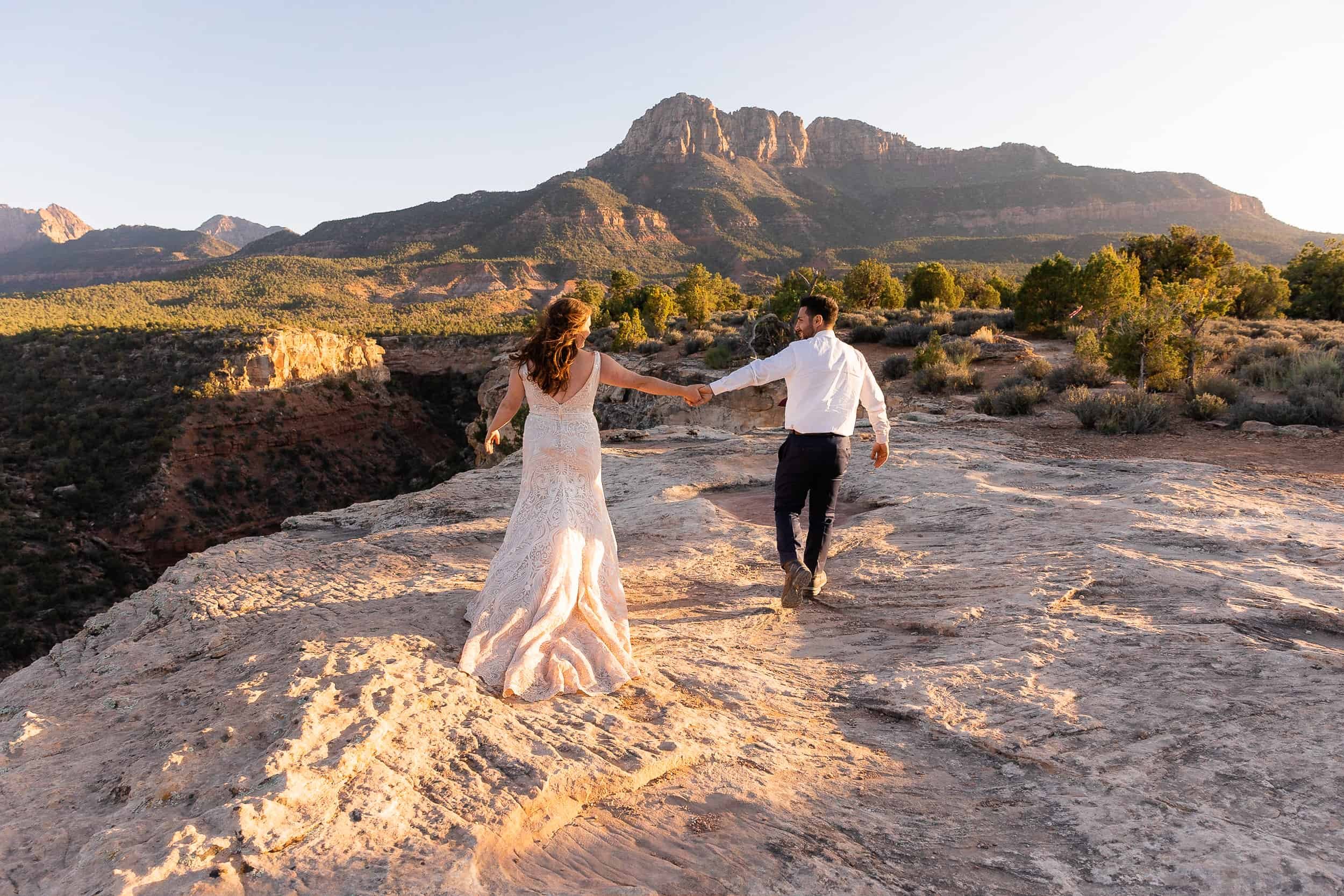 Bride and groom walk hand in hand in front of a red rock mountain peak during their elopement
