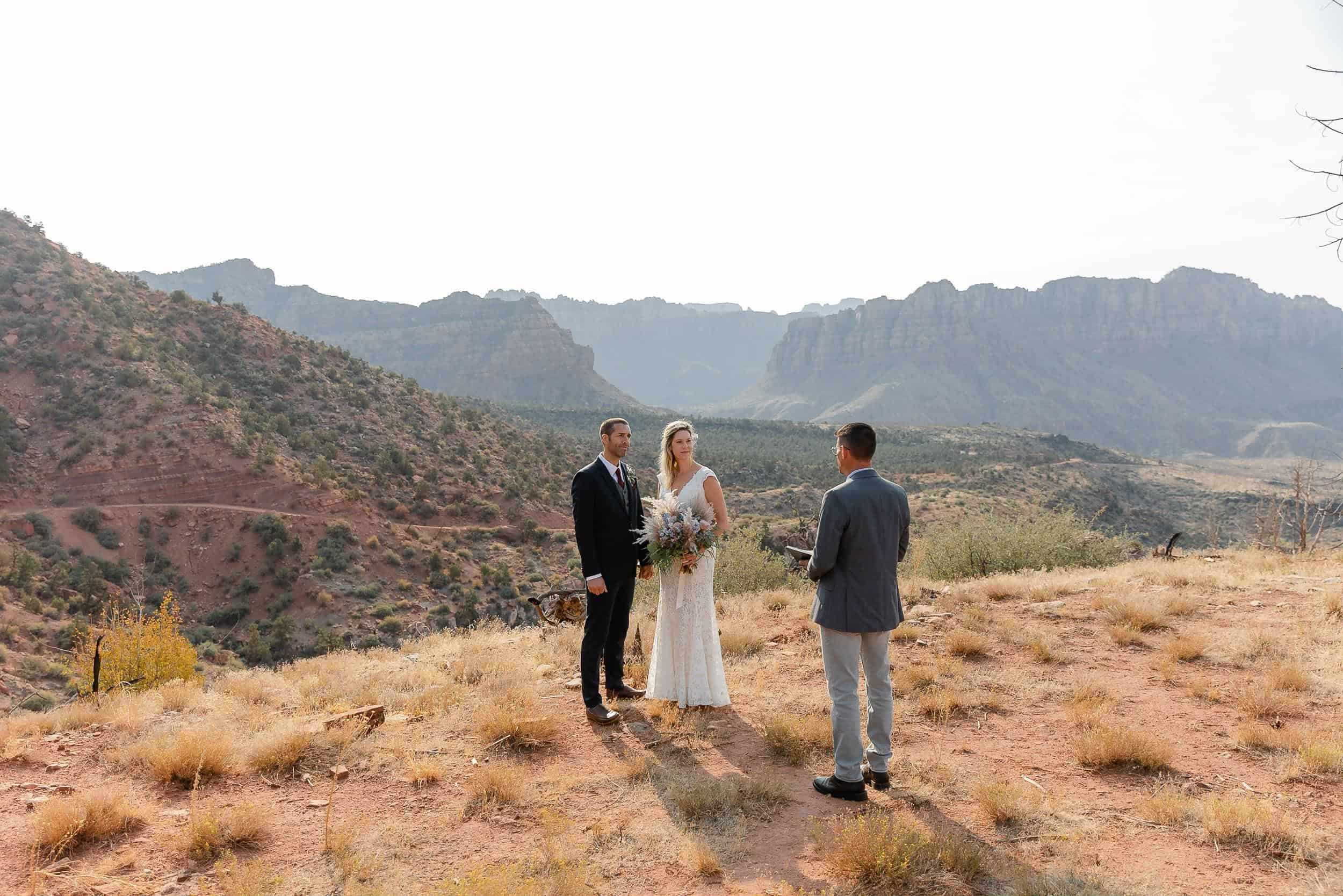 Couple stand together facing their officiant with a view of Zion National Park in the background at their elopement ceremony