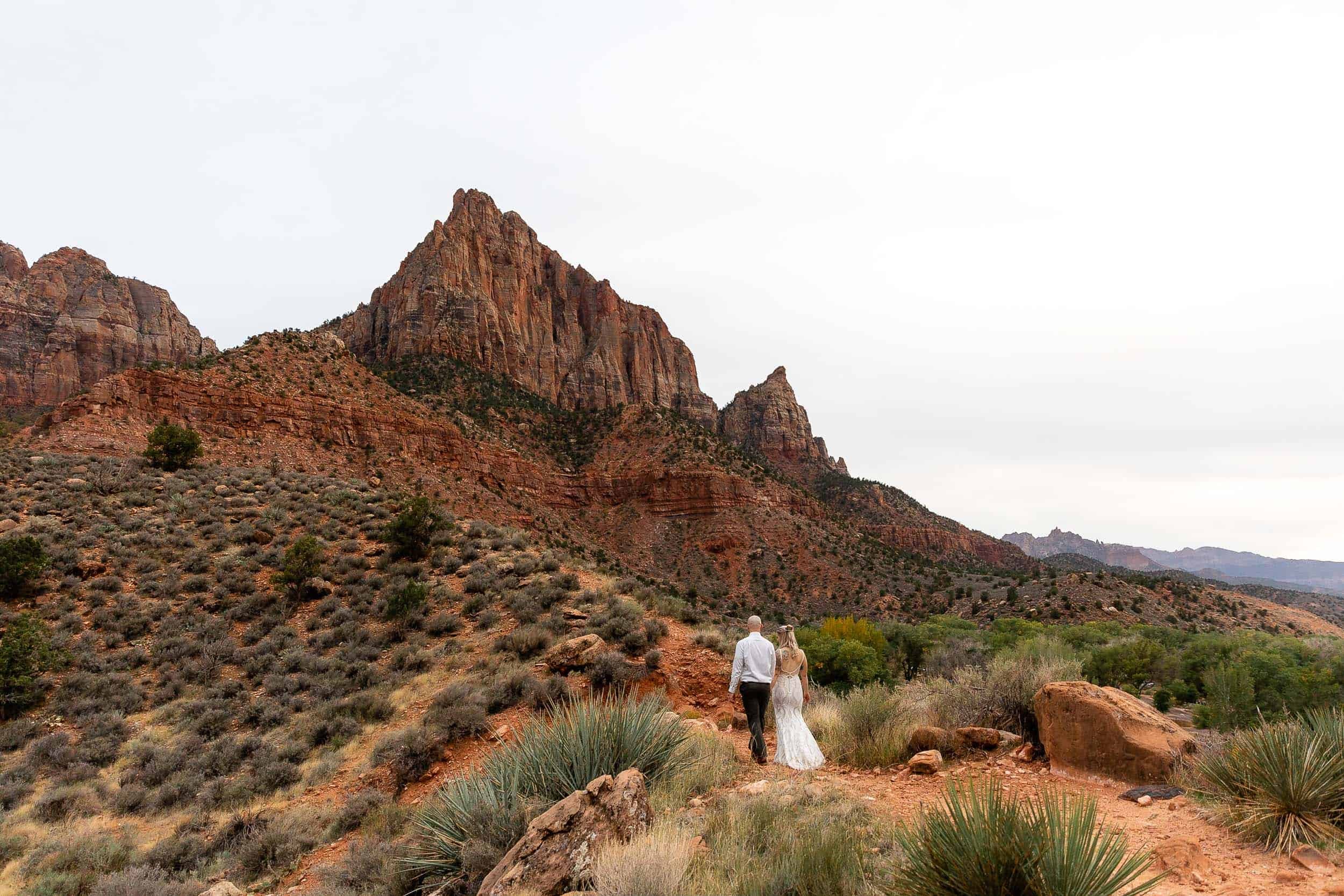 A couple walk along a path with the Watchman peak in Zion in the background
