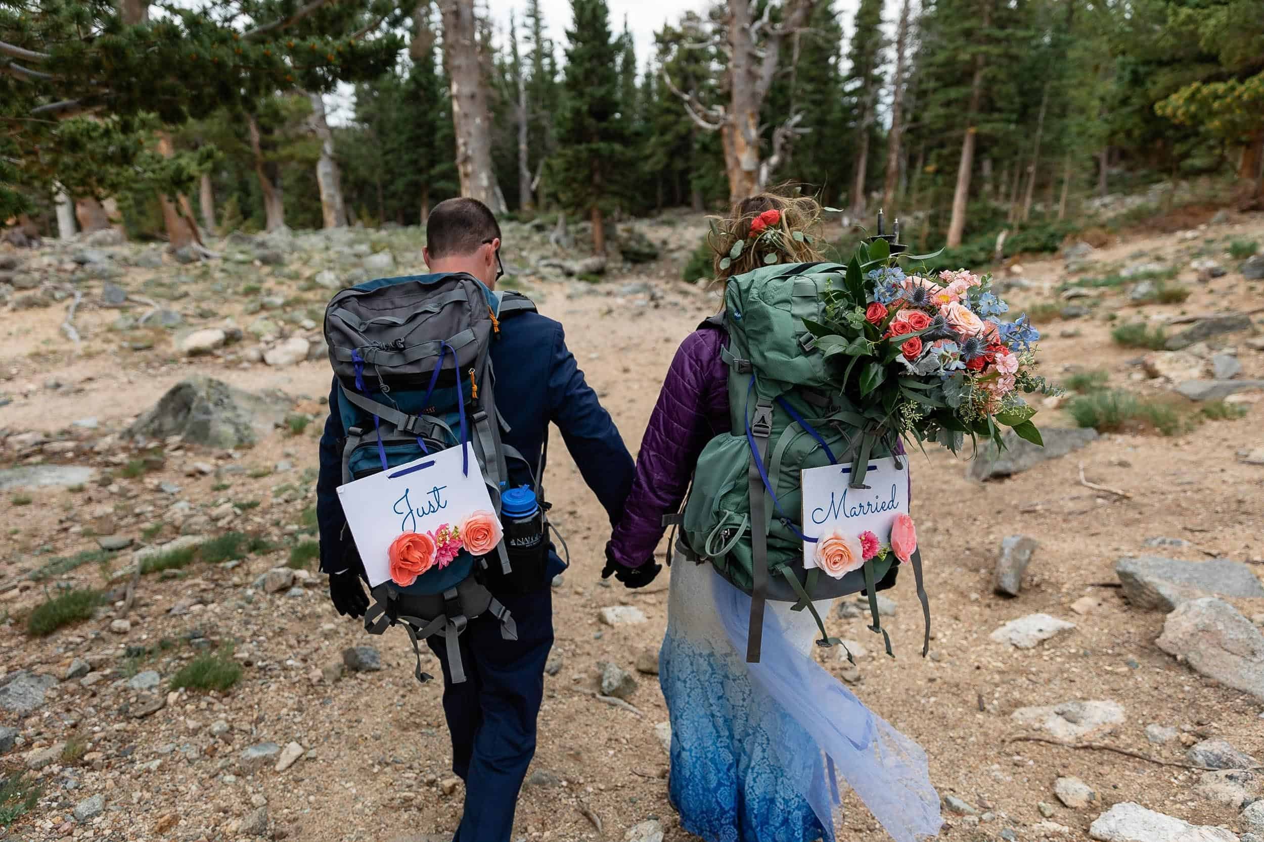Couple walk hand in hand away from the camera wearing backpacks with the signs reading just eloped on them