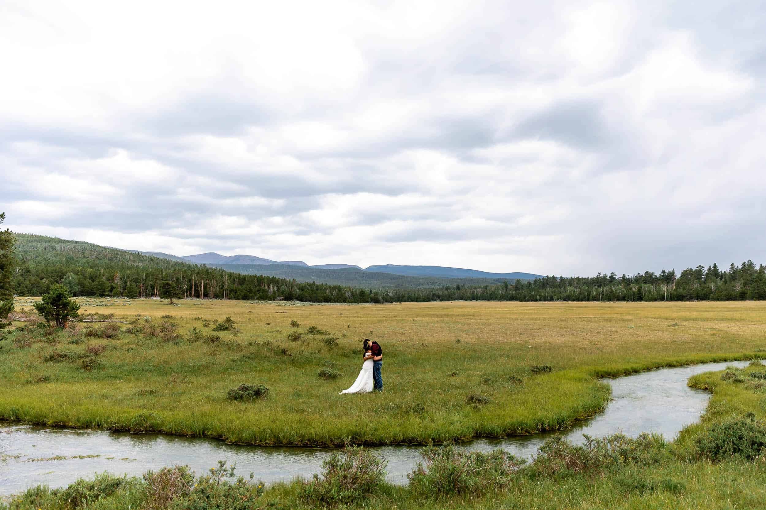 Wide angle shot of a couple embracing with a wide open mountain field and stream around them during their Utah elopement