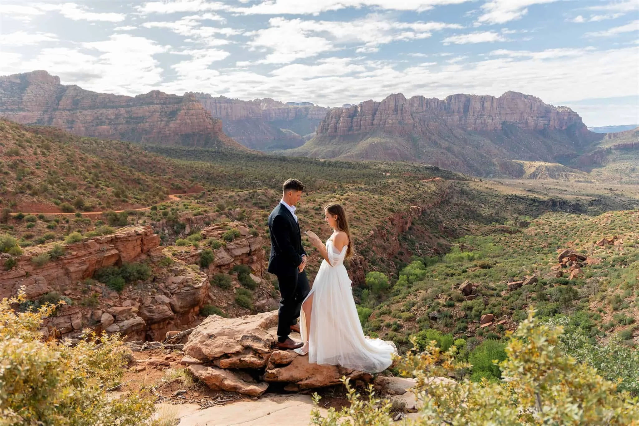 A couple is sharing vows outdoors in a desert landscape with red rocks and layered canyon cliffs in the background. The man is wearing a dark suit and the woman is wearing a white wedding dress, and they are looking at each other.