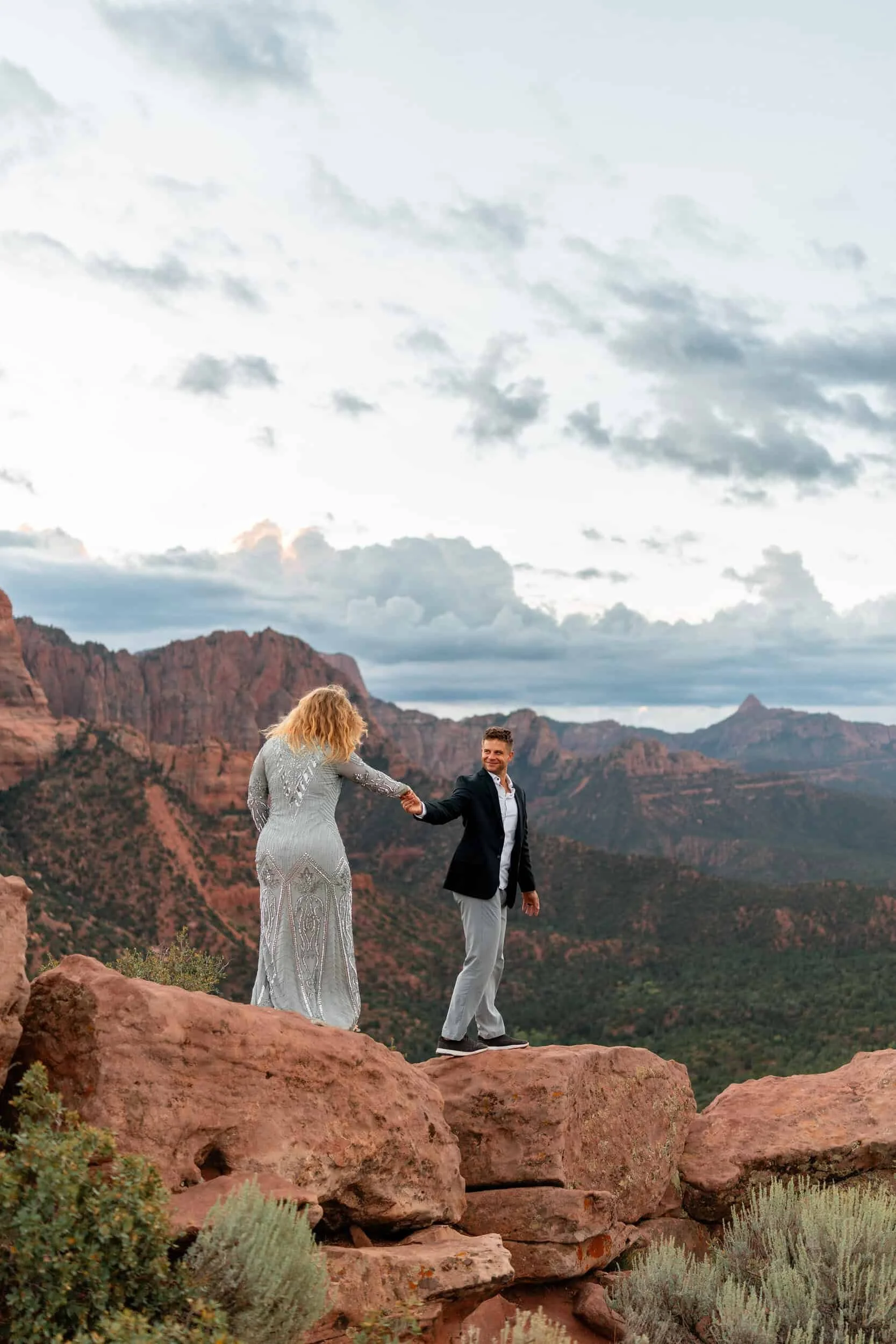 Couple in wedding clothes hold hands and walk on red rocks with the Kolob Canyons in the background at Timber Creek Overlook in Zion National Park