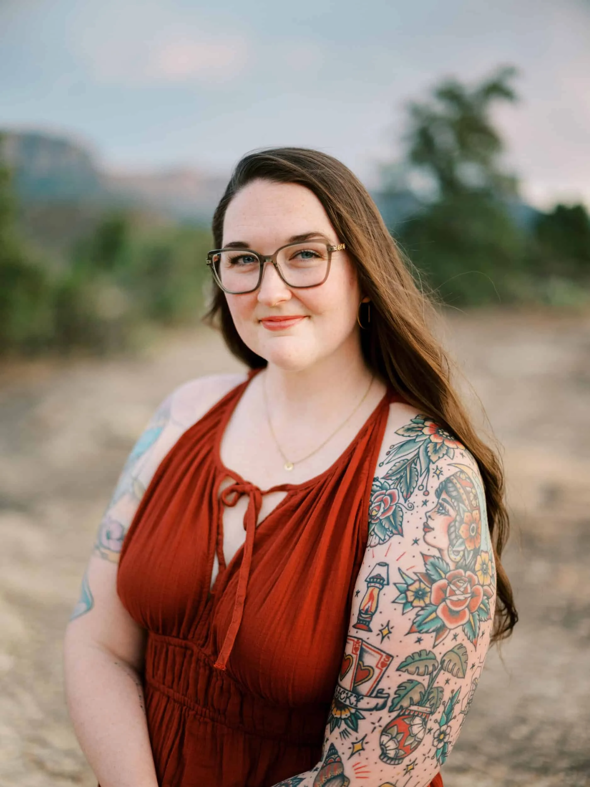 A woman with glasses and long brown hair, wearing a rust-colored sleeveless dress, smiling outdoors. She has colorful tattoos on her left arm, including a woman’s face, flowers, a lighthouse, and stars.