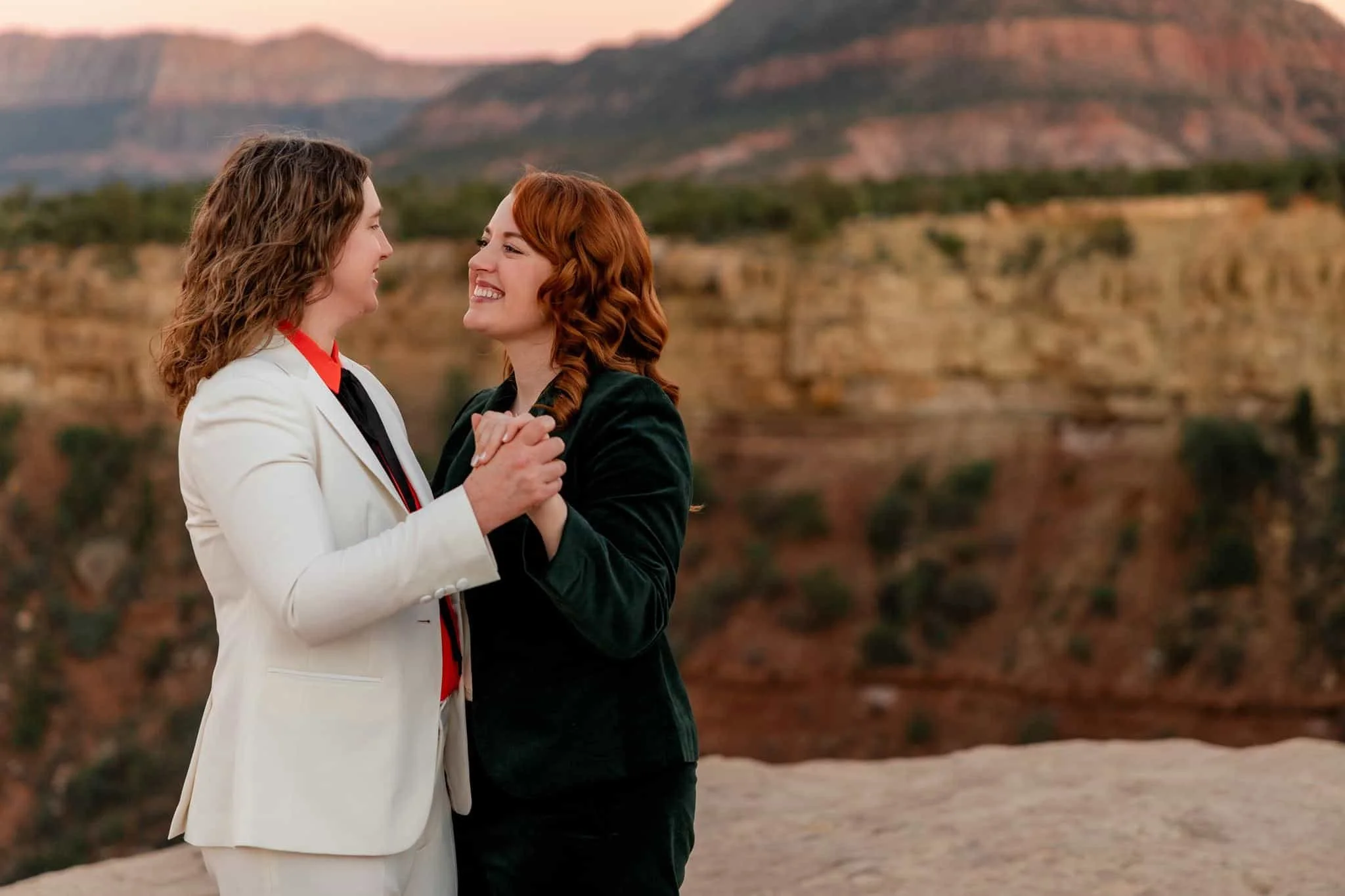 Two women holding hands and smiling at each other and dancing outdoors with mountains in the background during sunset during their lgbtq zion wedding