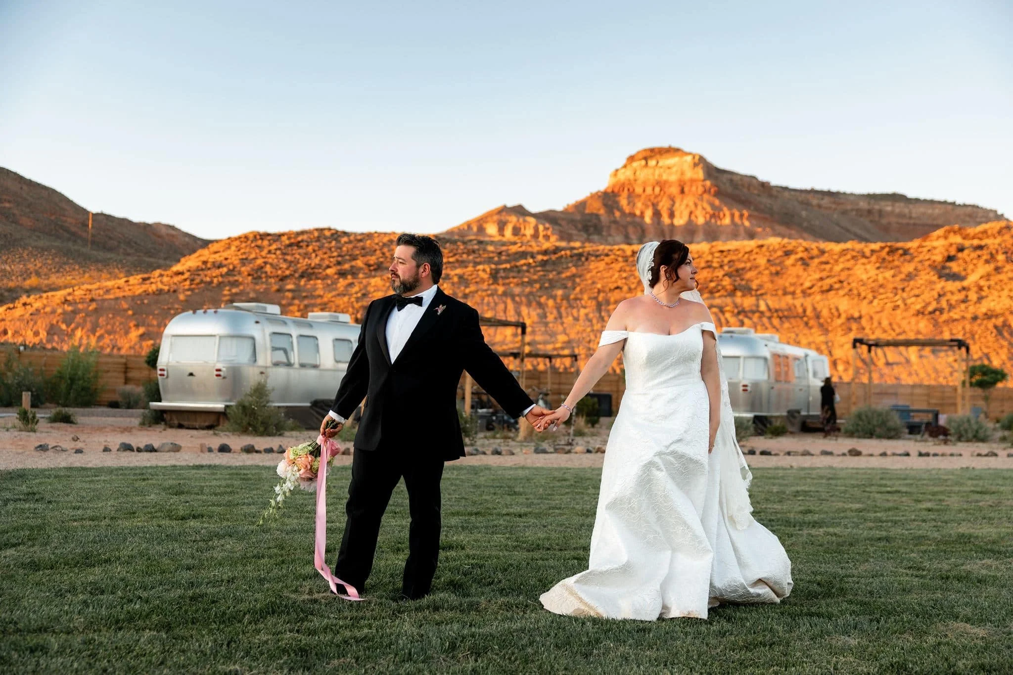 Bride and groom stand in a green lawn holding hands with Airstreams and red mountains lit up by sunset during their wedding at AutoCamp Zion