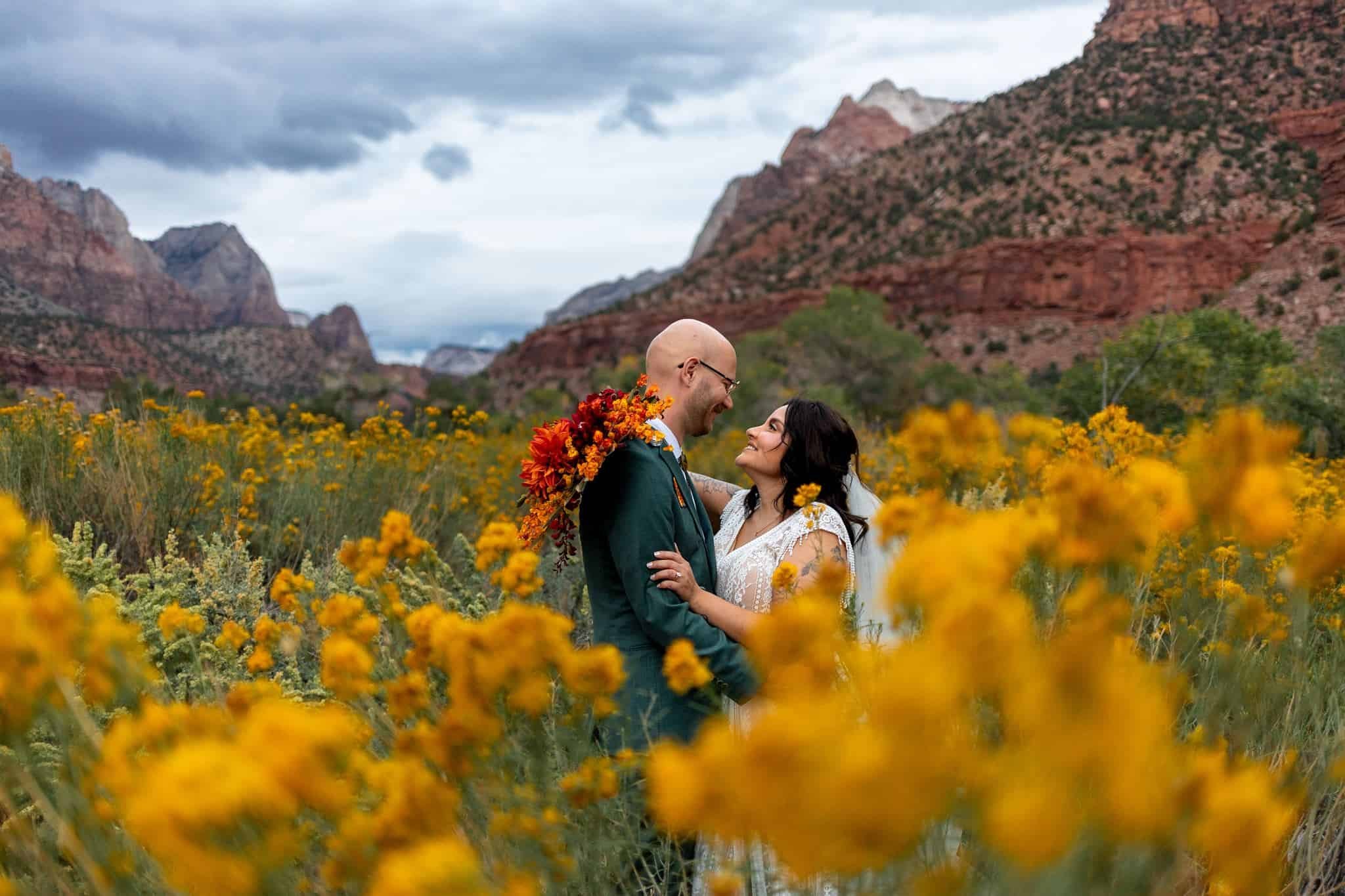 A man and woman in a wedding photo standing in a field of yellow flowers with red rocky mountains in the background.