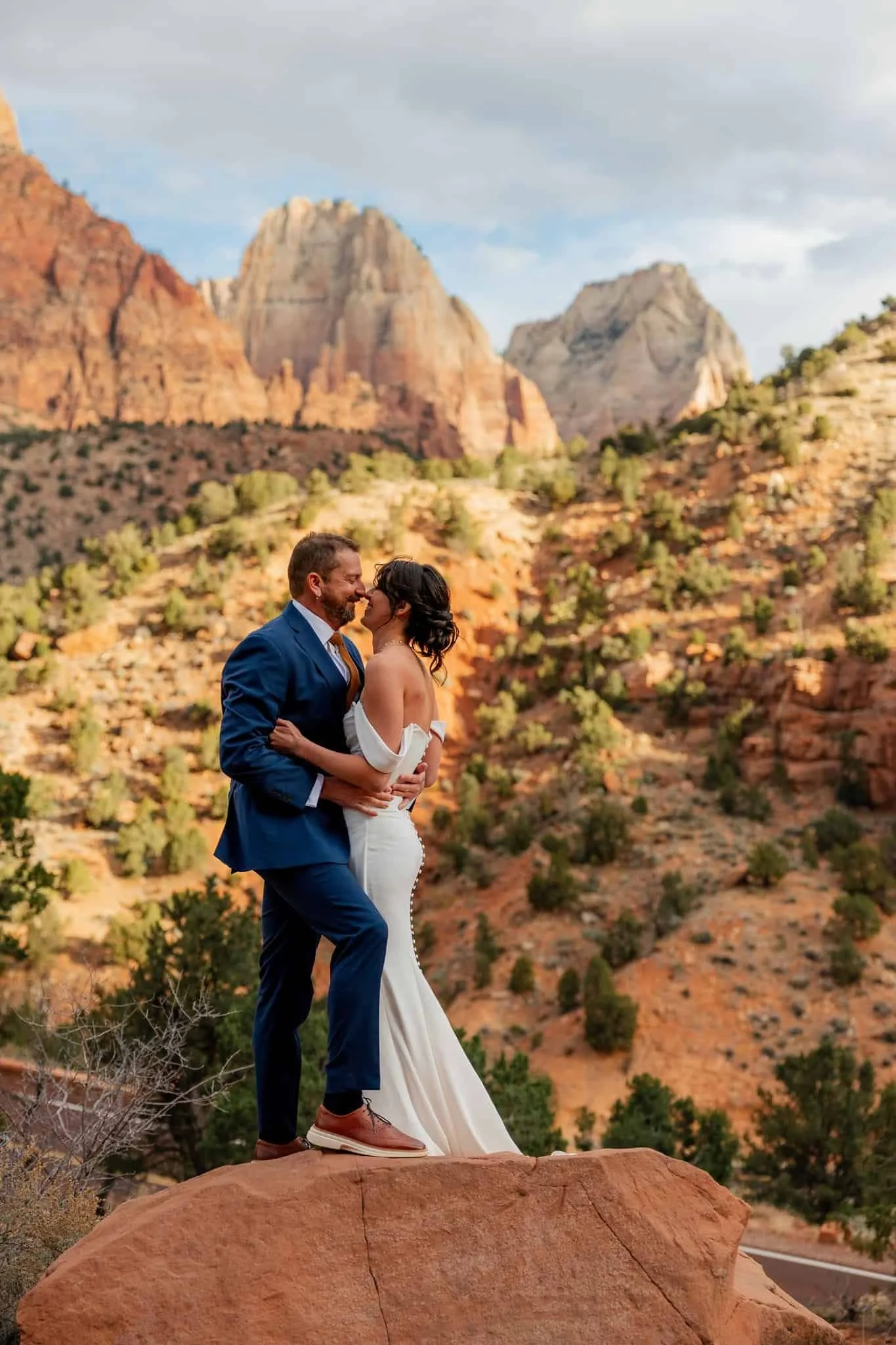 A couple dressed in wedding attire standing on a rocky outcrop in a desert landscape with red rocks and mountains in the background, sharing an intimate moment in Zion National Park