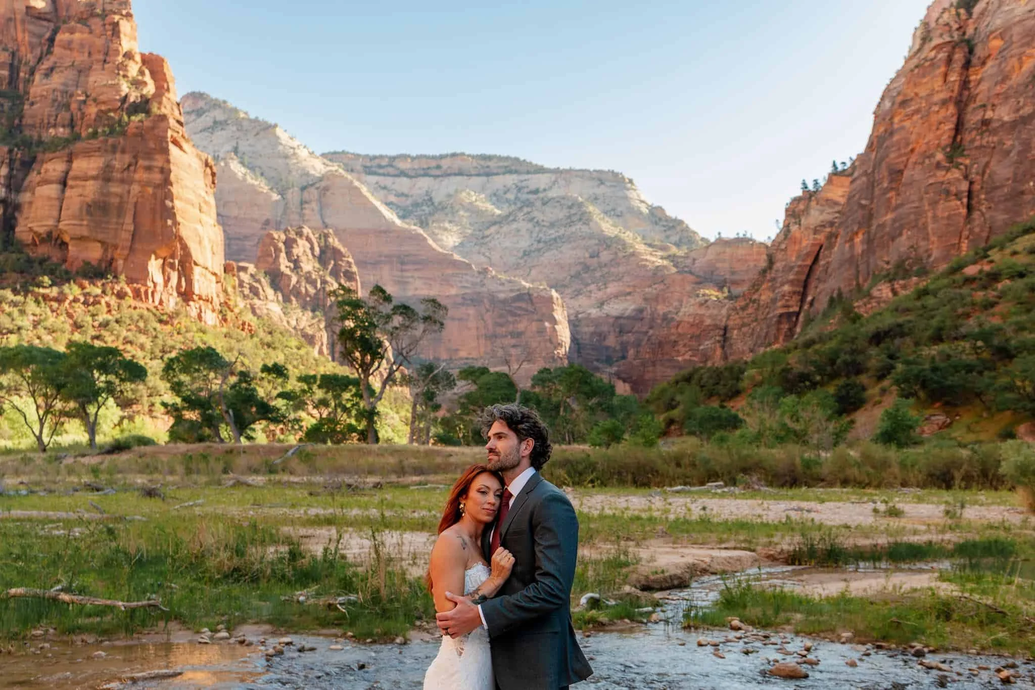 A man and a woman standing closely together in a grassy area next to the Virgin River in Zion National Park, with a large canyon with red and white rock formations and green trees in the background during daytime.