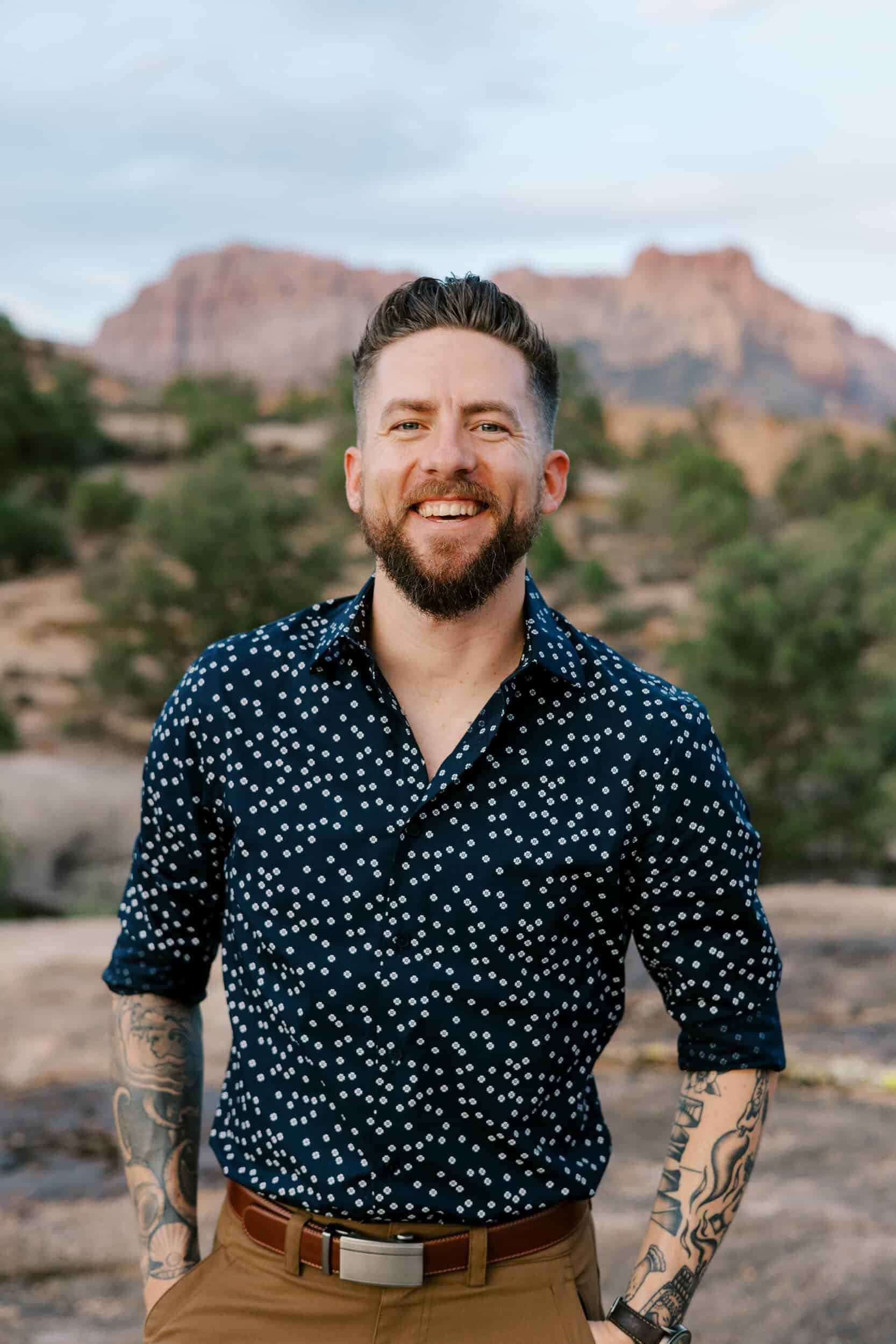 Man with tattoos wearing a navy blue shirt with white polka dots, khaki pants, and a brown belt, standing outdoors with desert mountains in the background.