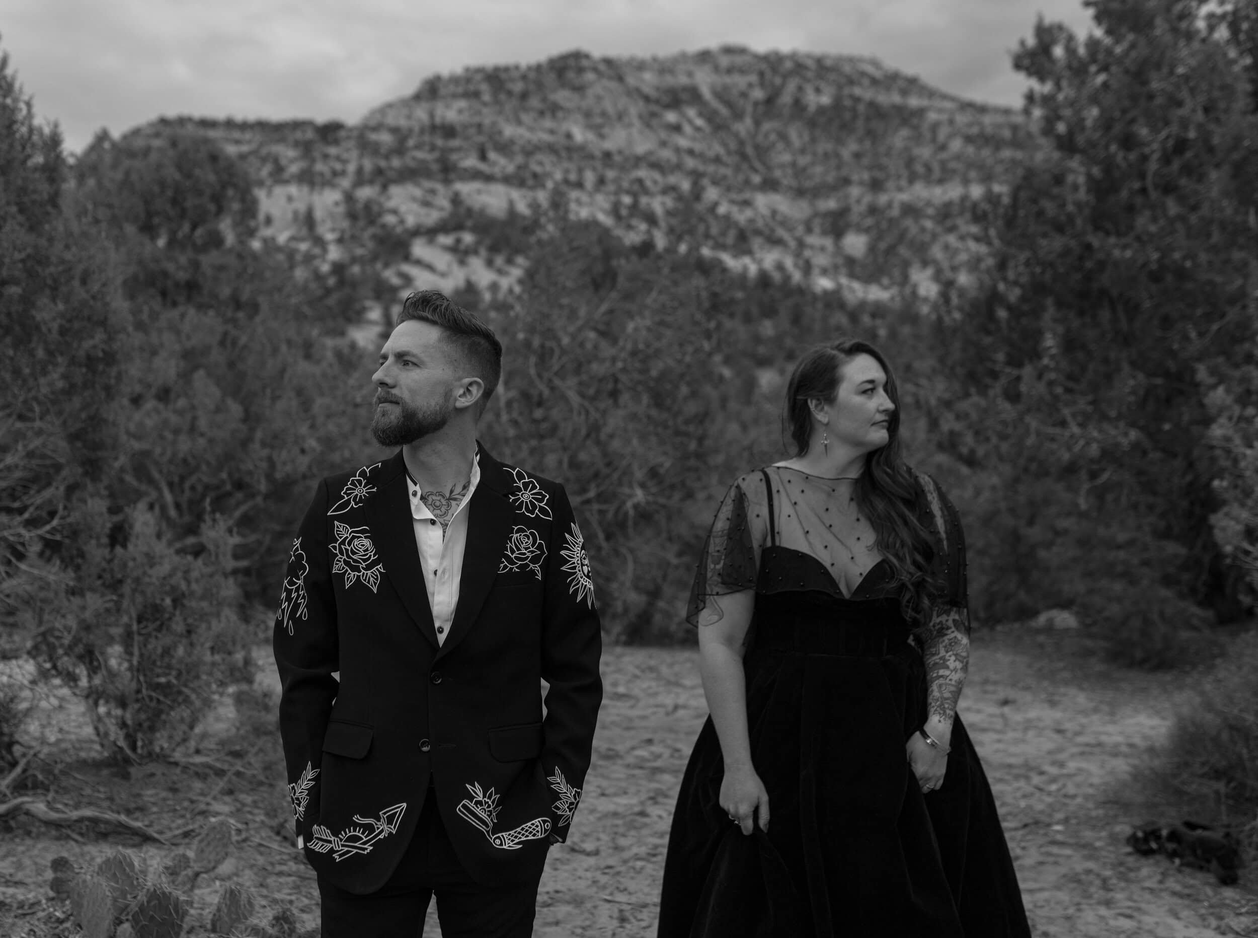 Black and white photo of a man and woman standing outdoors with mountains and trees in the background. The man has a beard and styled hair, wearing a dark suit jacket with embroidered floral patterns over a white shirt. The woman has long hair, wearing a sheer polka-dot top with lace detailing, and is looking to the side.