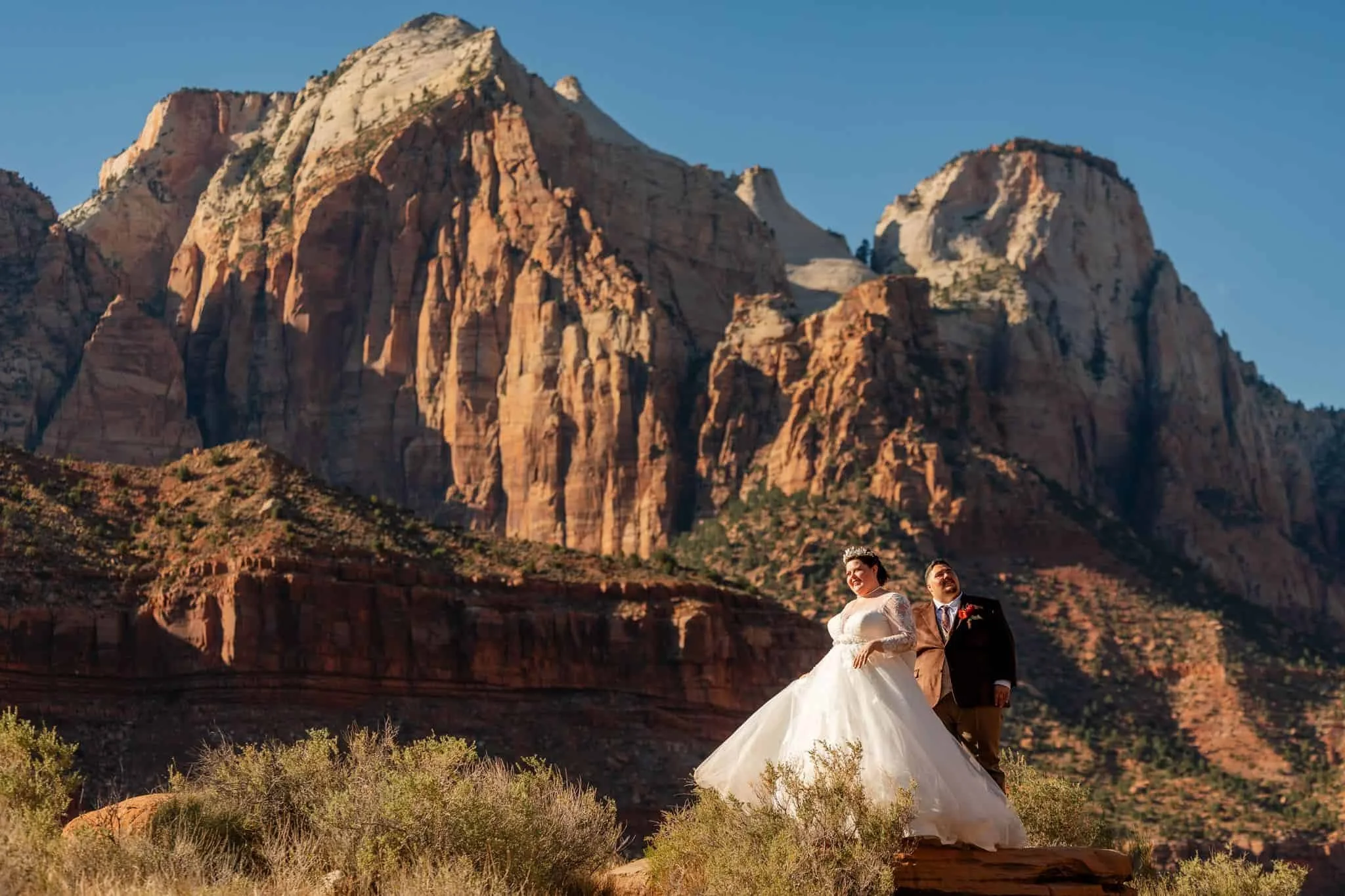 A plus sized bride and groom walking in a desert landscape with red rock formations and mountains in the background in Zion National Park