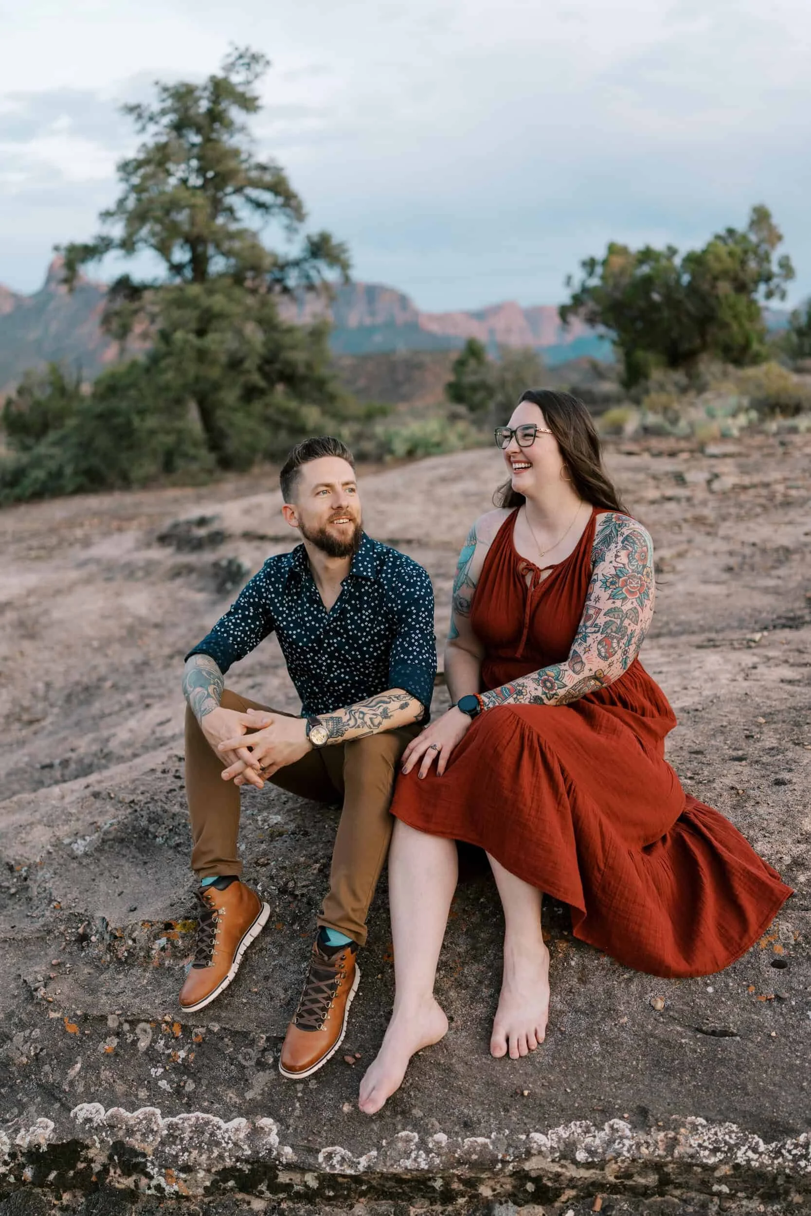 A man and woman sitting on a rock outdoors with trees and distant mountains in the background, laughing and smiling, dressed casually, with tattoos on their arms.