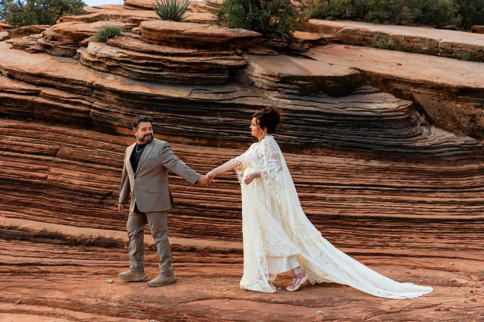 A couple dressed in formal attire holding hands and walking in front of layered red rock formations in a desert landscape during sunset in Zion