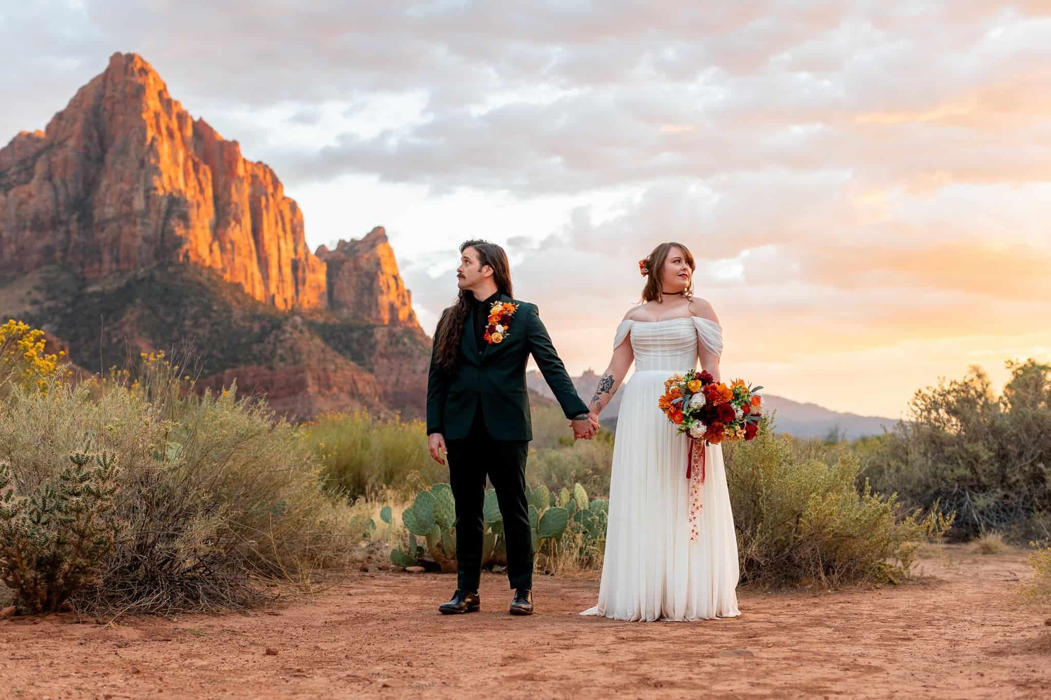 A couple holding hands in a desert landscape with mountains and clouds in the background, one dressed in a dark suit with floral accents and the other in a white off-shoulder dress with a bouquet of flowers during their zion wedding