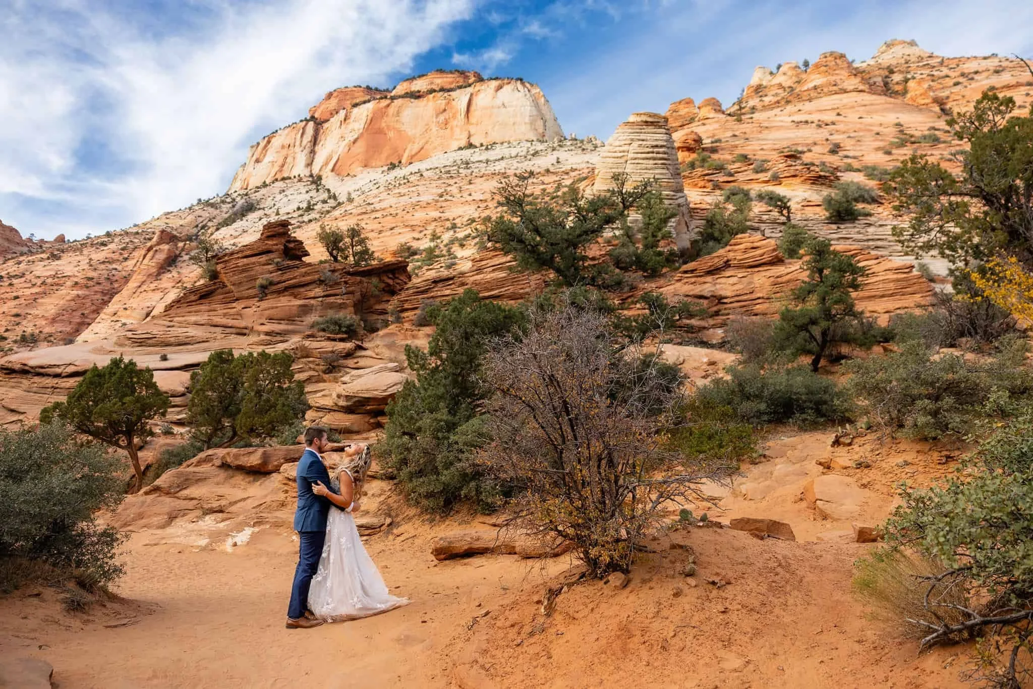 A bride and groom kissing outdoors in a desert landscape with layered red and white rock formations and sparse trees.